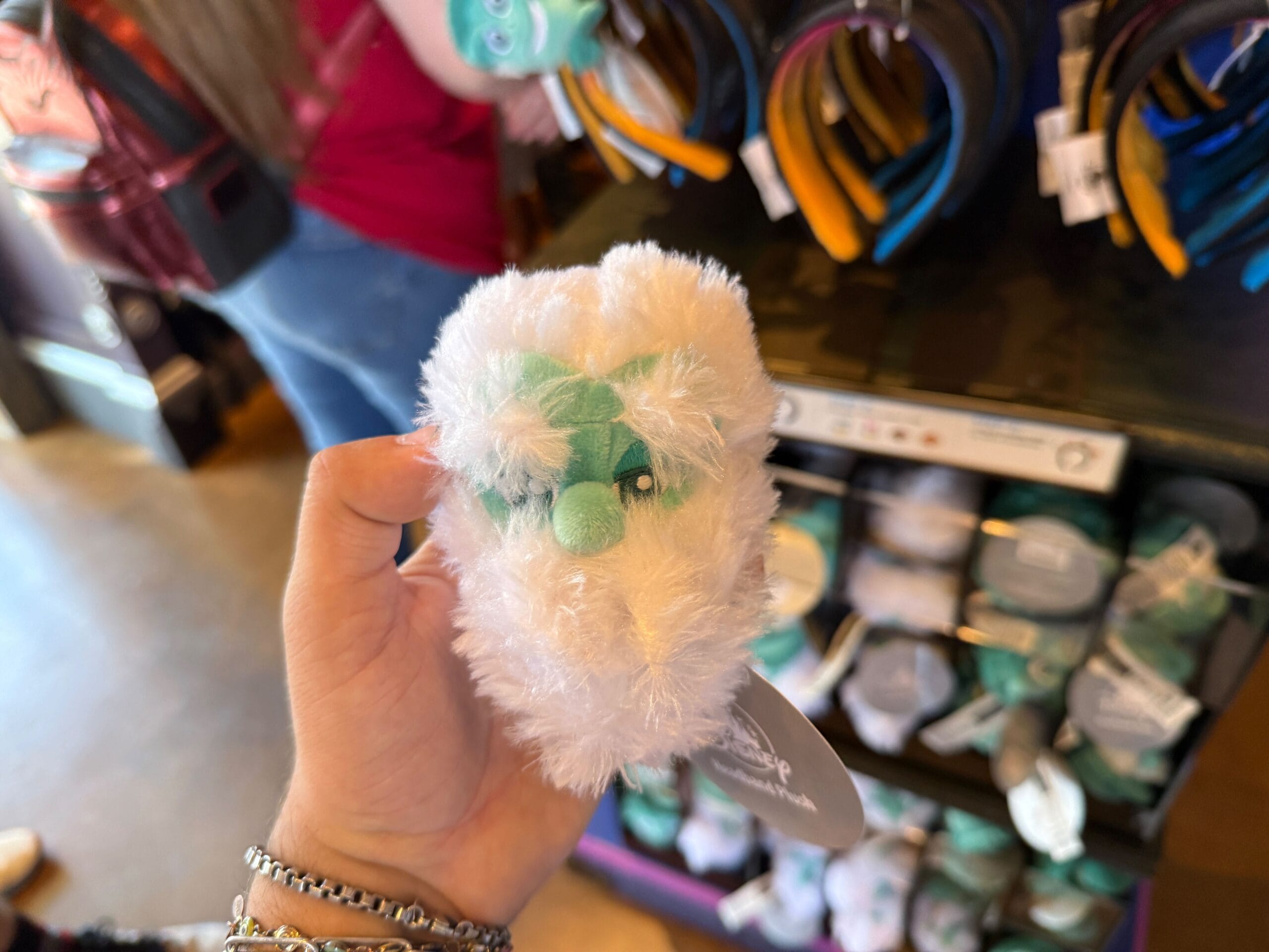 A hand holds a small, fluffy white plush toy with green details from the HM Merchandise at Disneyland.
