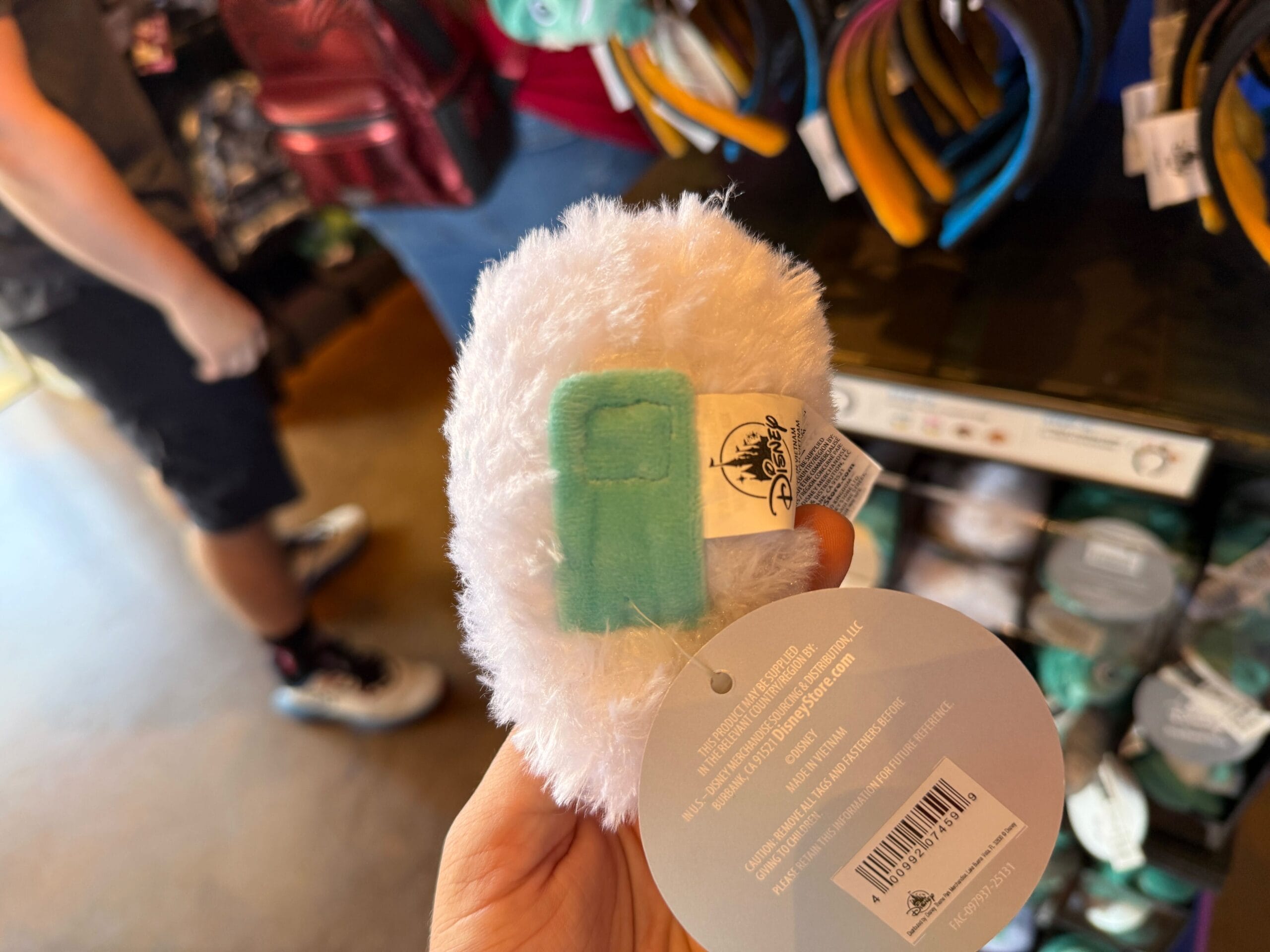 A hand holds a fluffy white Disneyland keychain with a teal patch in an HM Merchandise store display, tags visible.