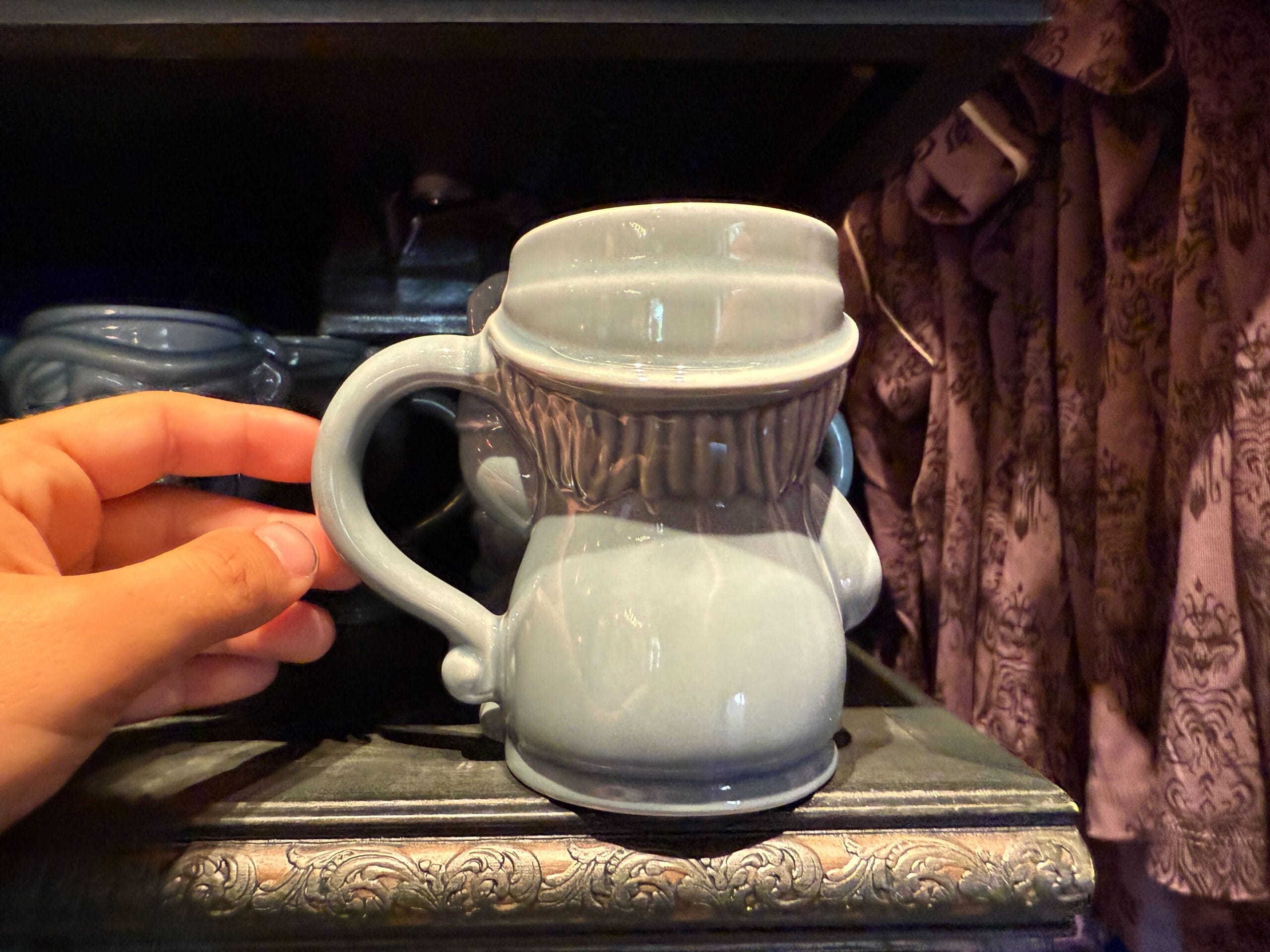 A hand holds a gray HM Merchandise mug with a wavy shape, displayed on a shelf next to patterned purple fabric.
