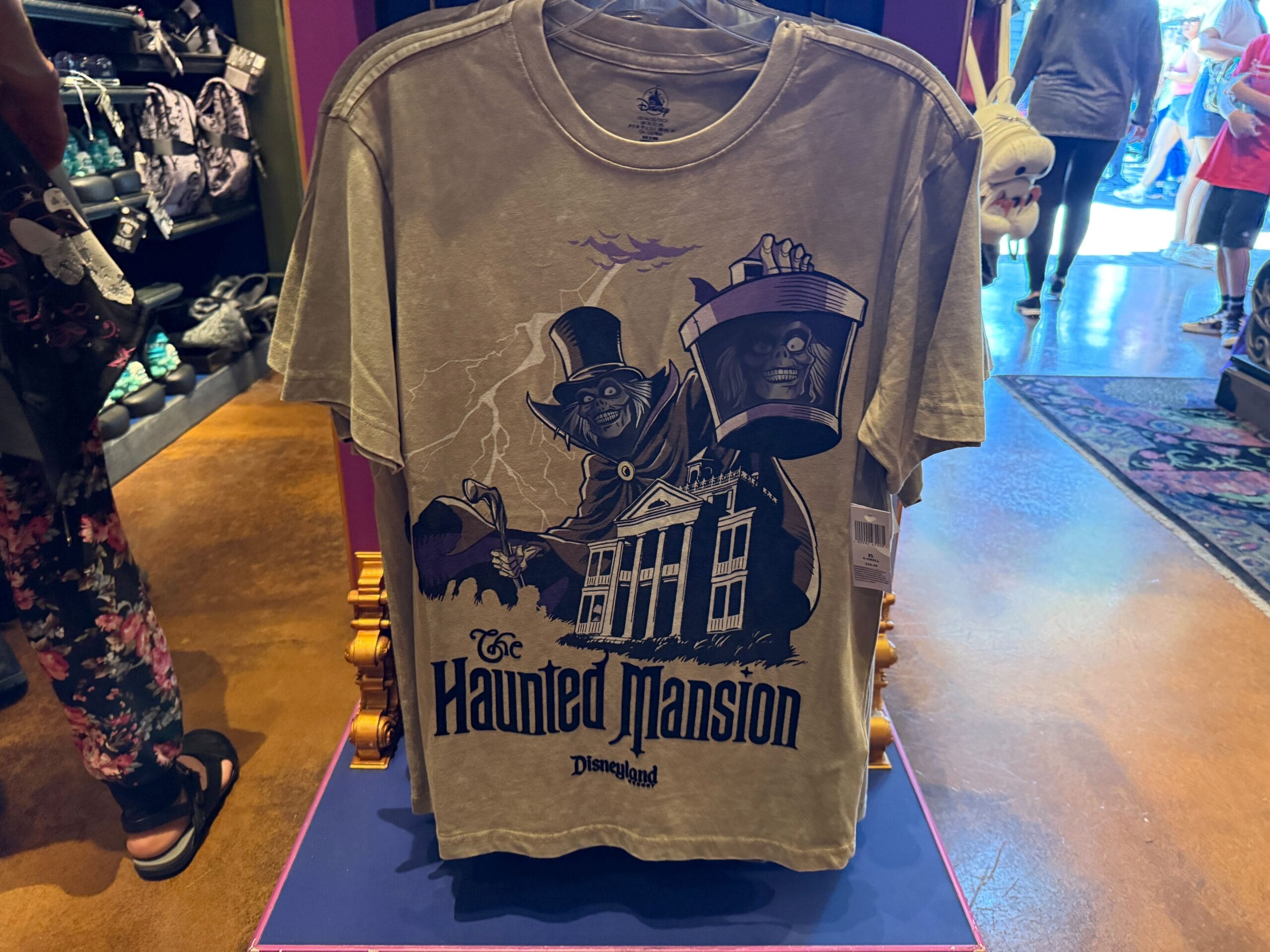 A gray HM Merchandise T-shirt featuring the Hatbox Ghost and Haunted Mansion building on display in Disneyland.