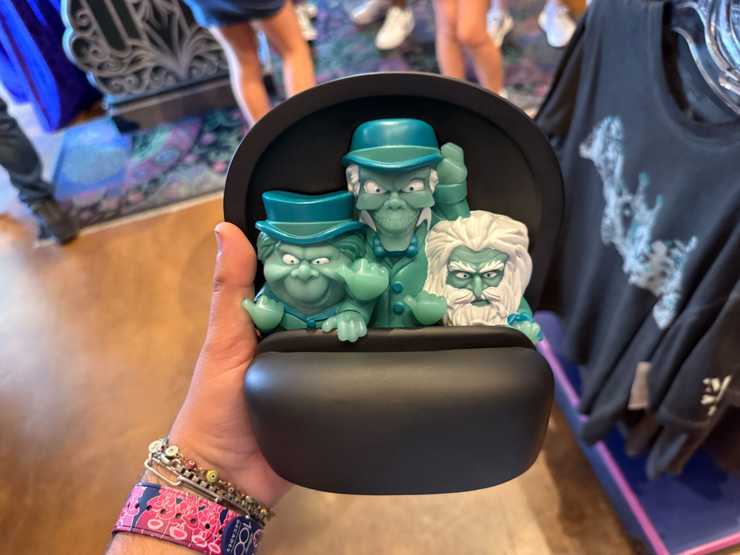 A hand holds a hatbox with three blue-green ghost figurines—Disneyland HM Merchandise and people in the background.