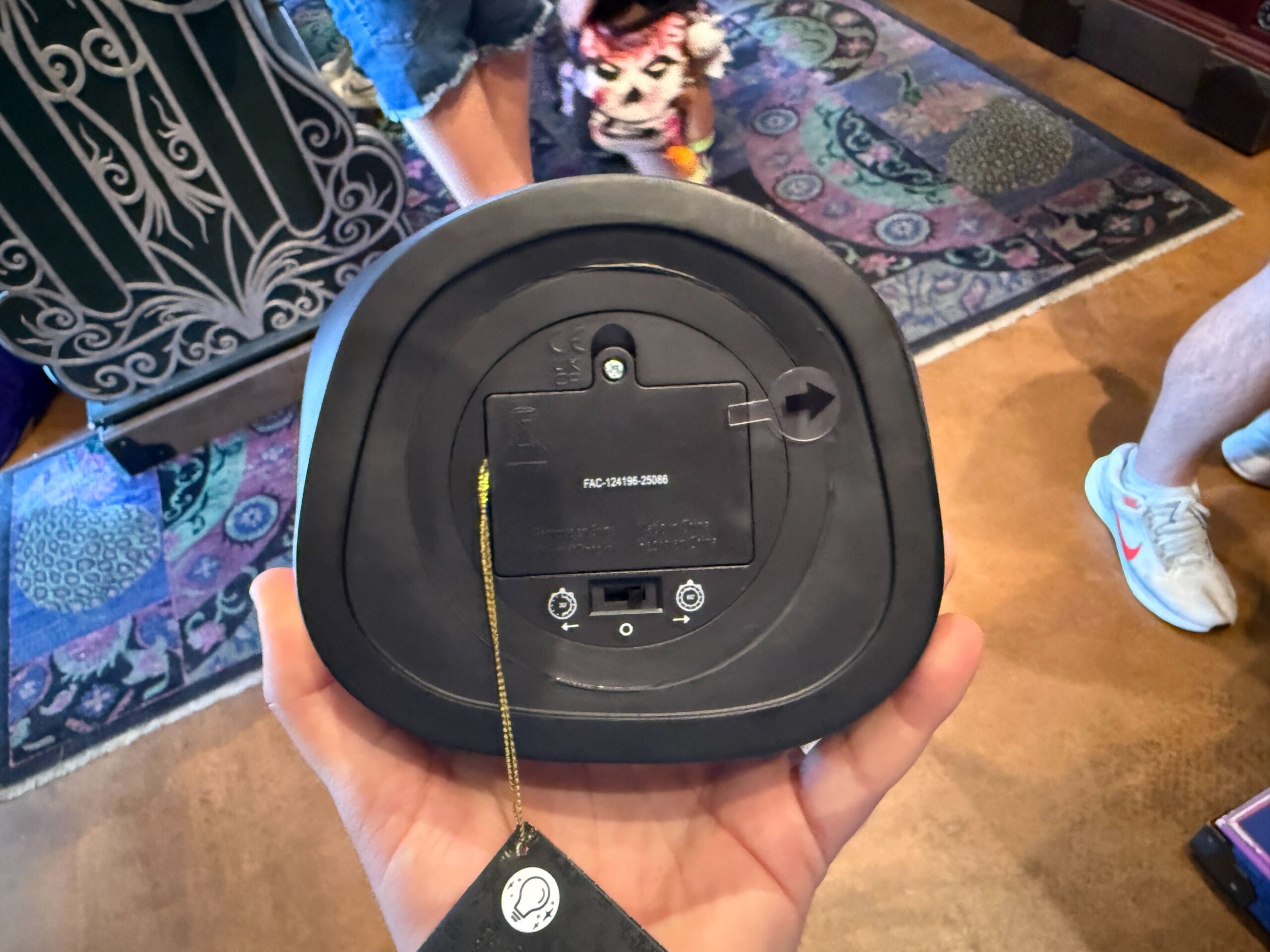 A hand holds a black electronic device upside down, showing its HM Merchandise battery compartment and barcode.