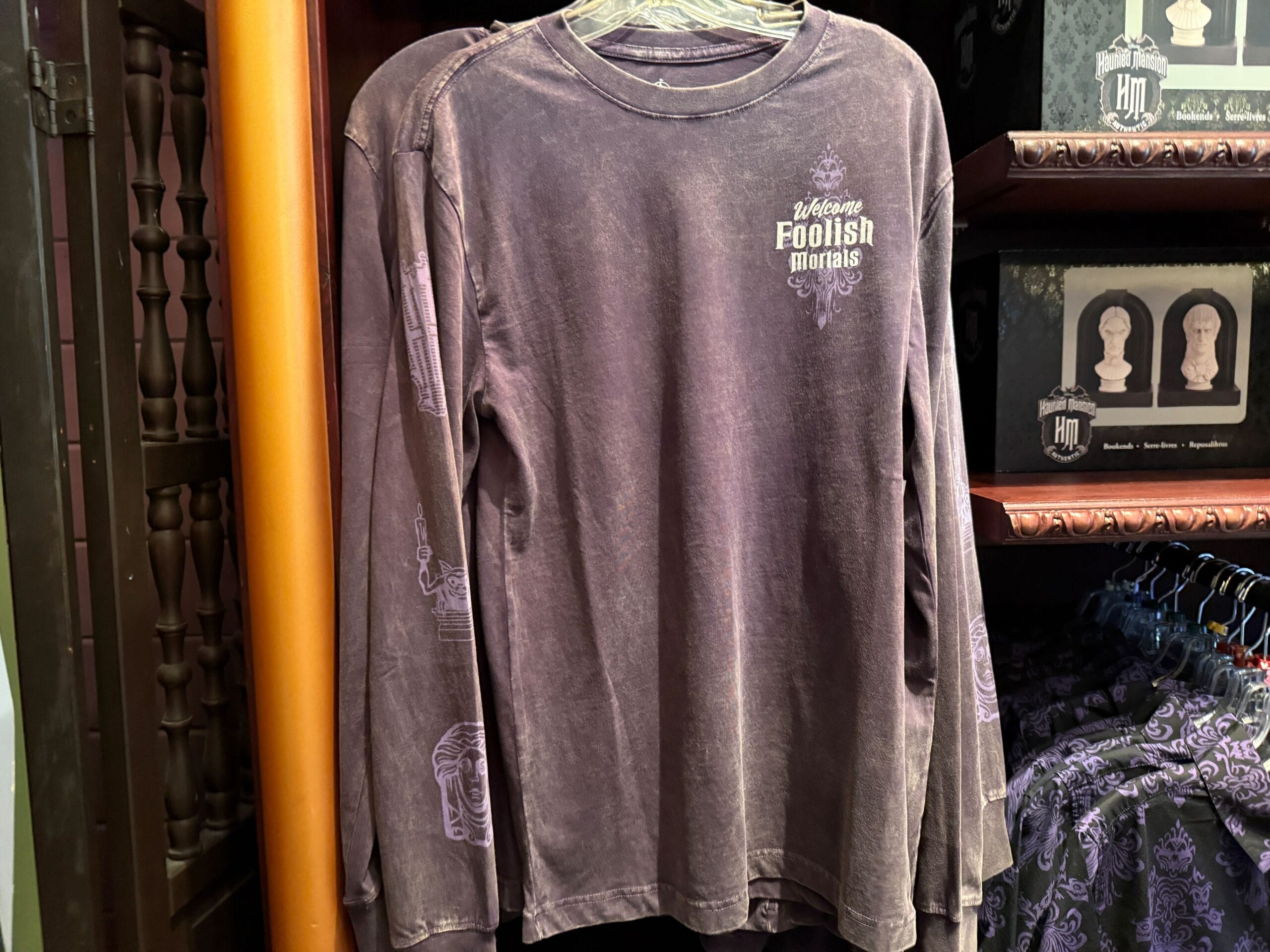 A long-sleeve shirt with "Welcome, Foolish Mortals" text displayed on a rack of HM Merchandise.