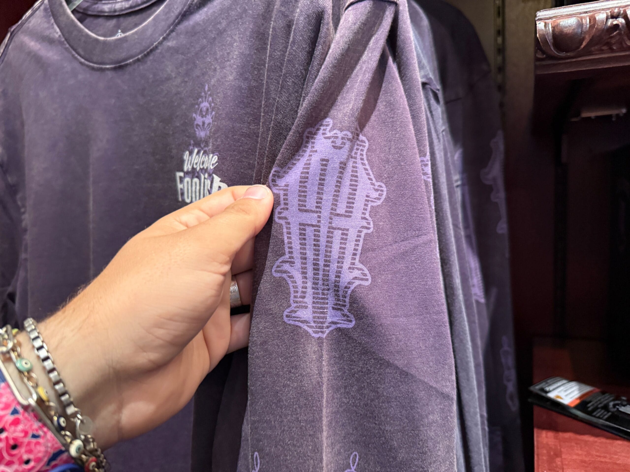 A hand holds a purple HM Merchandise shirt with a Haunted Mansion logo and "Where Frights" text in white and lavender.