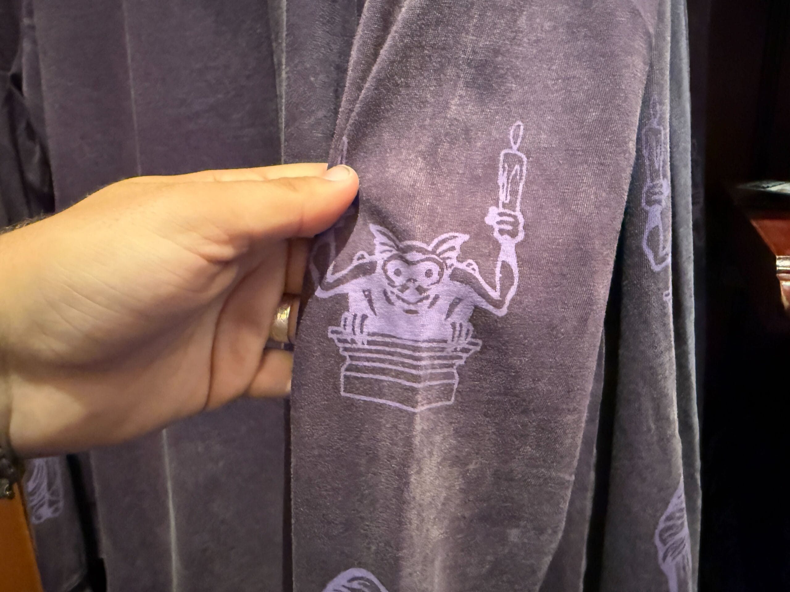 A hand holds purple fabric featuring a cartoonish gargoyle holding a candle, inspired by Disneyland HM Merchandise.