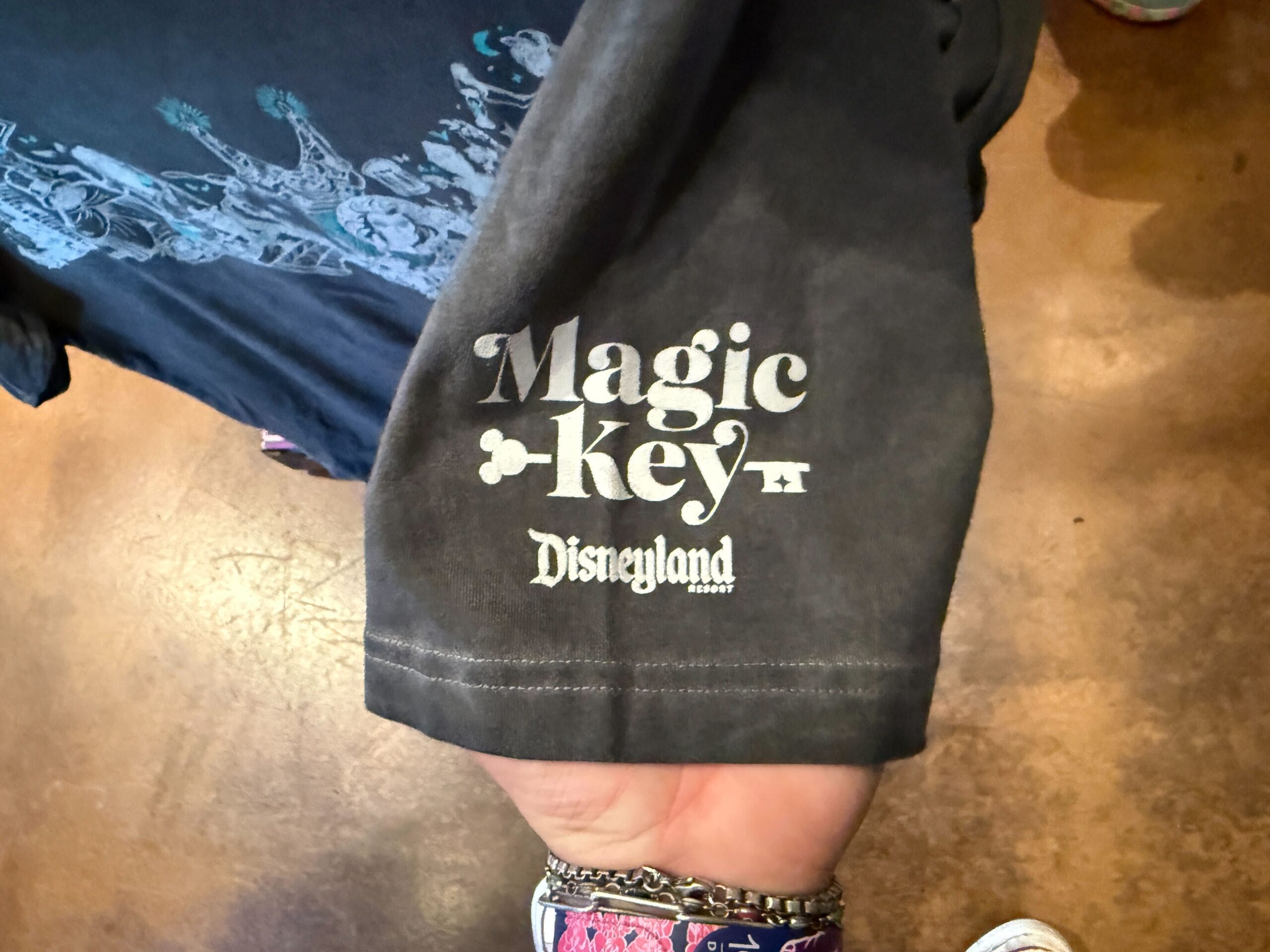A hand holds a black HM Merchandise shirt with "Magic Key Disneyland Resort" printed in white on the sleeve.