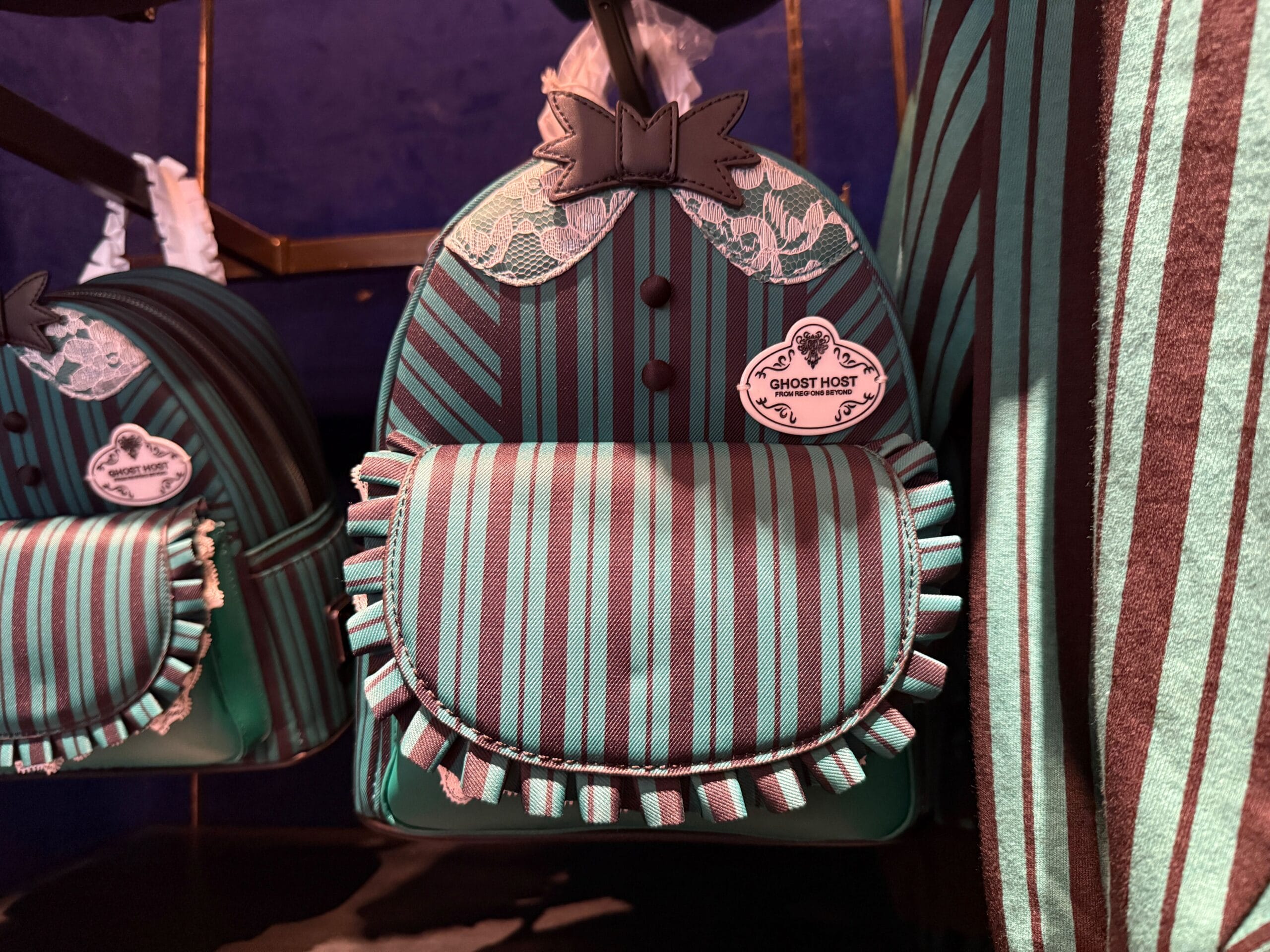 A teal and black striped "Ghost Host" backpack with ruffles, lace, and HM Merchandise badge on display at Disneyland.