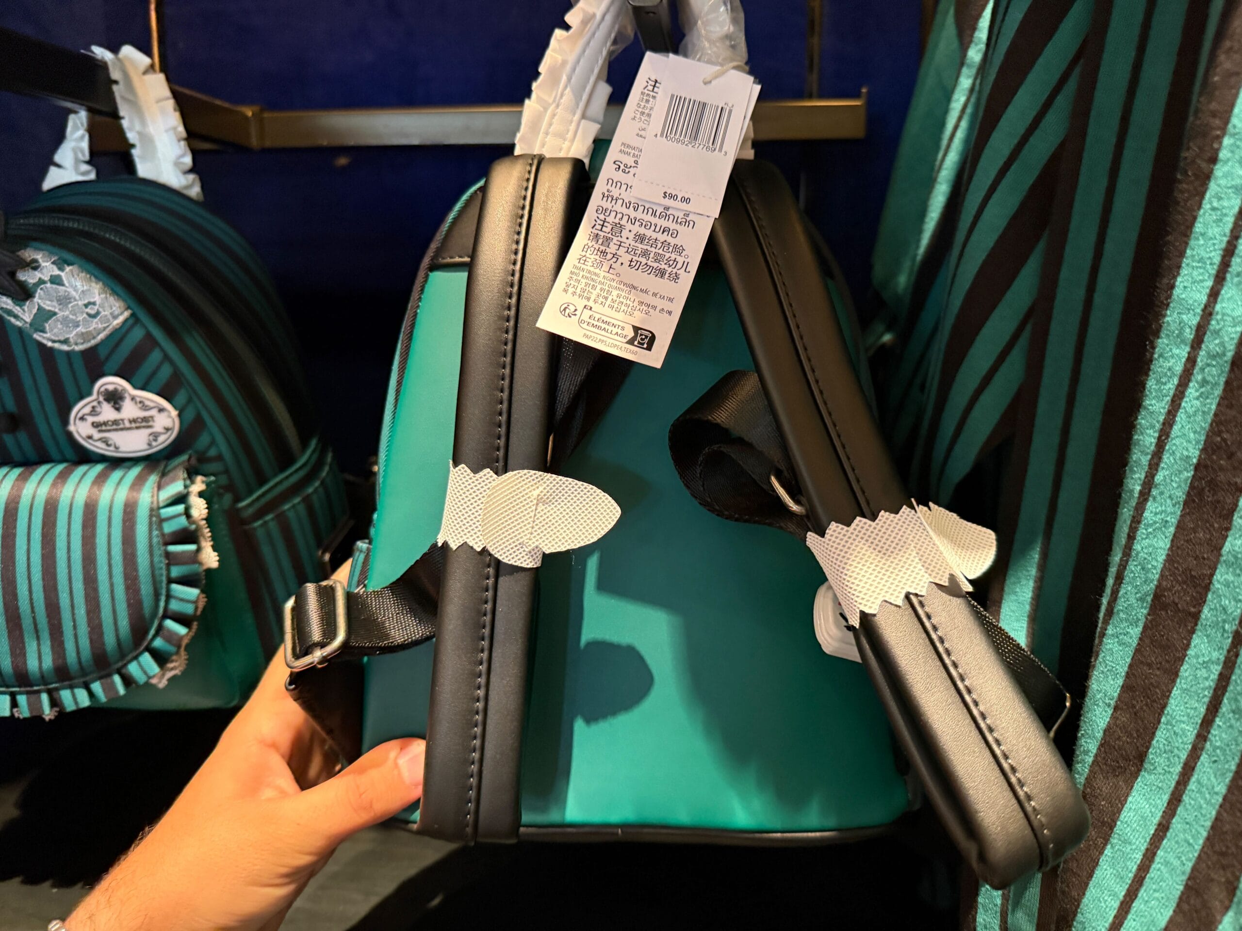 A hand holds a teal and black striped backpack with bat-shaped zipper pulls from Disneyland's HM Merchandise display.