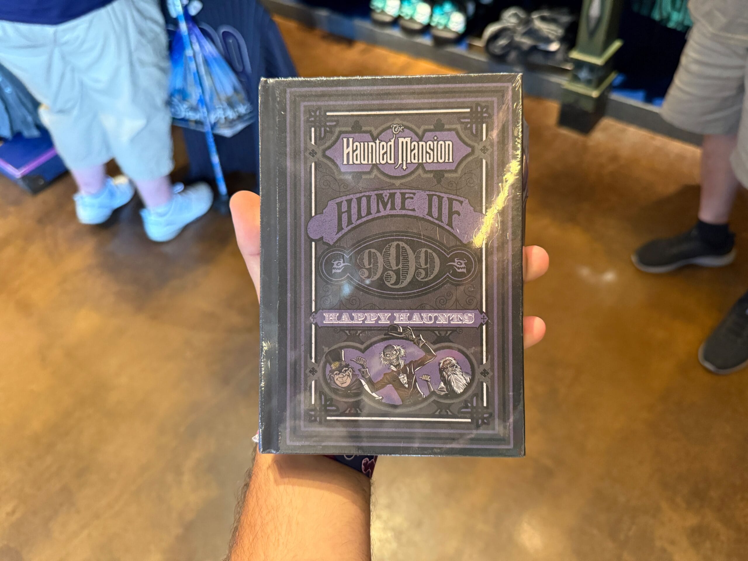 A person holds Disneyland HM Merchandise—a Haunted Mansion book with "Home of 999 Happy Haunts" on the cover—in a store.