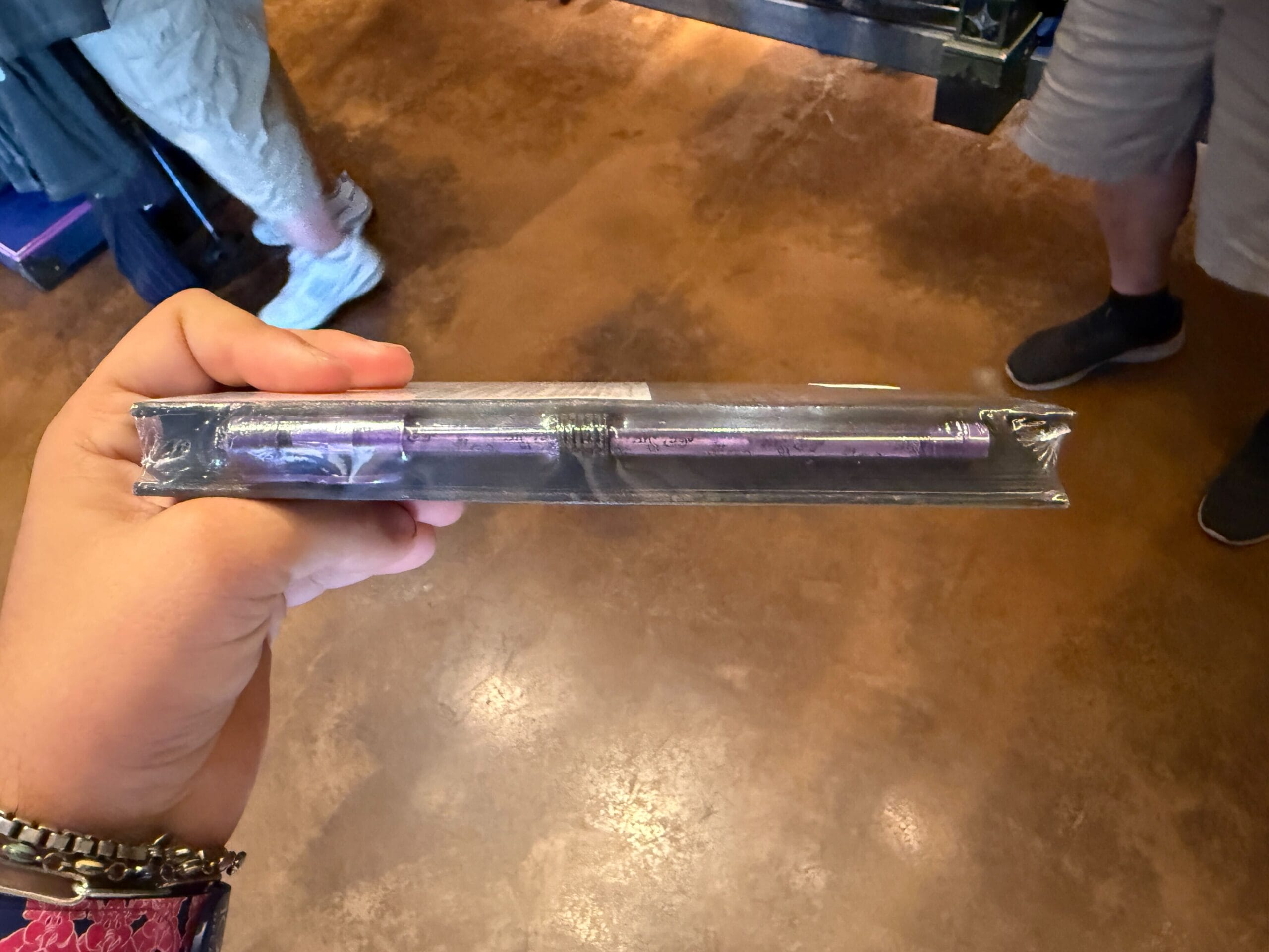 A hand holds a sealed HM Merchandise item, possibly a book, with two pens on a brown floor background.