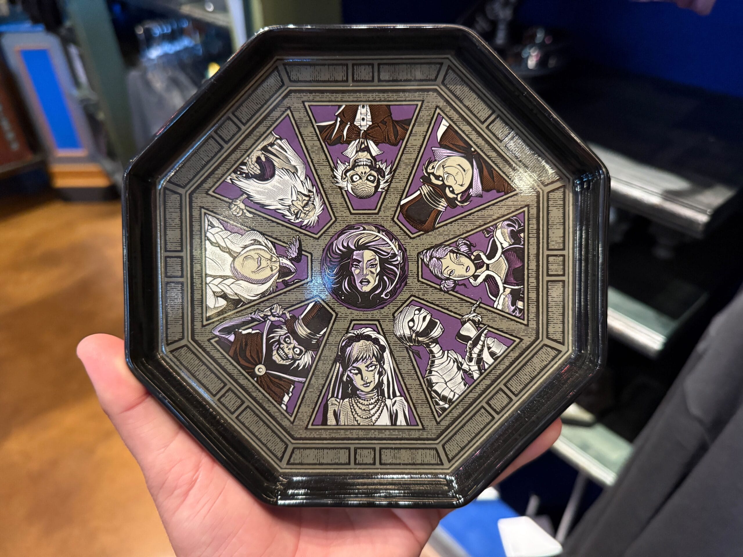 A hand holds an octagonal plate featuring HM Merchandise with illustrated spooky characters on a dark, decorative background.