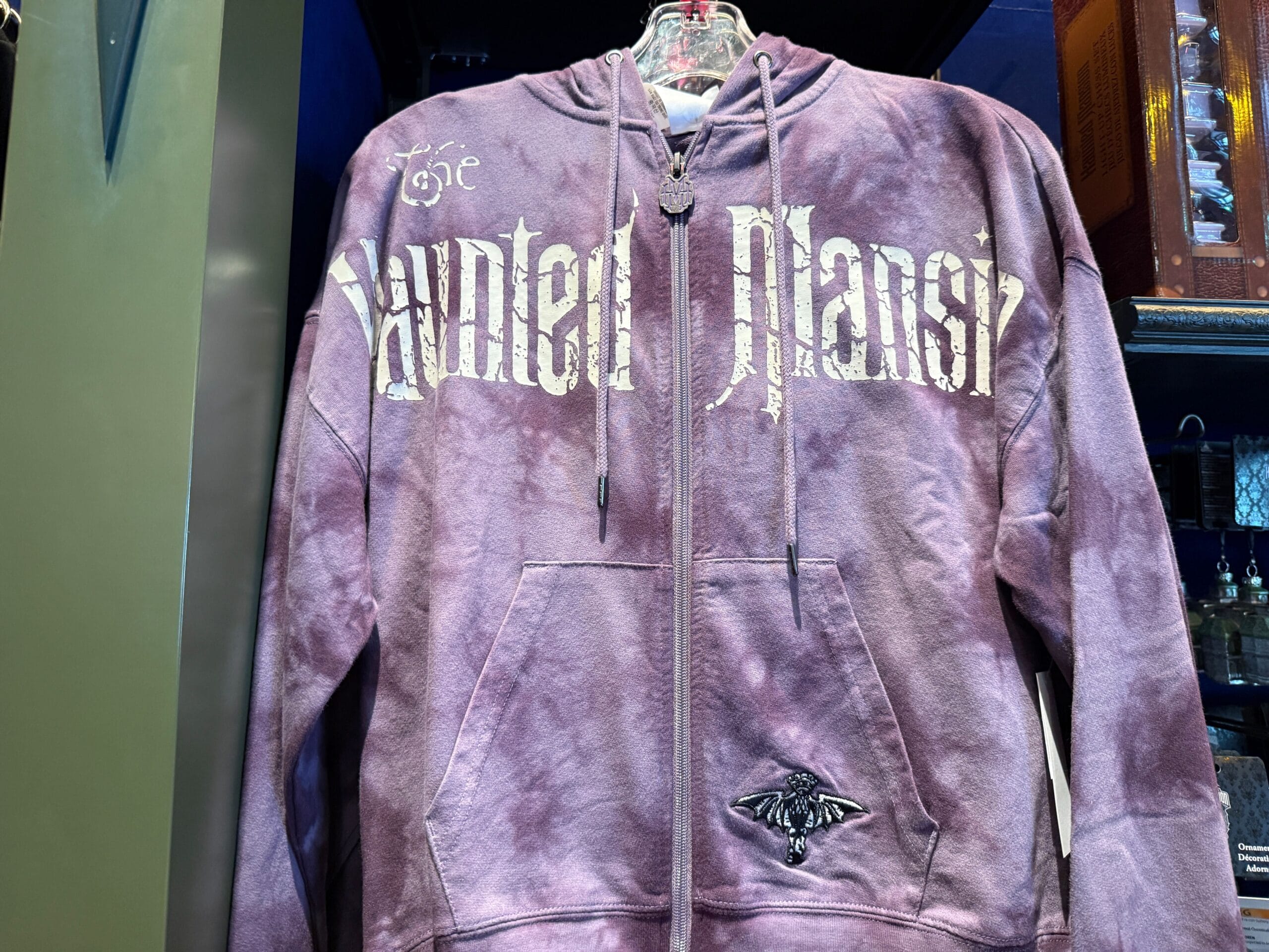 Purple tie-dye hoodie with "The Haunted Mansion" text, small moth graphic, and classic HM Merchandise at Disneyland on display.