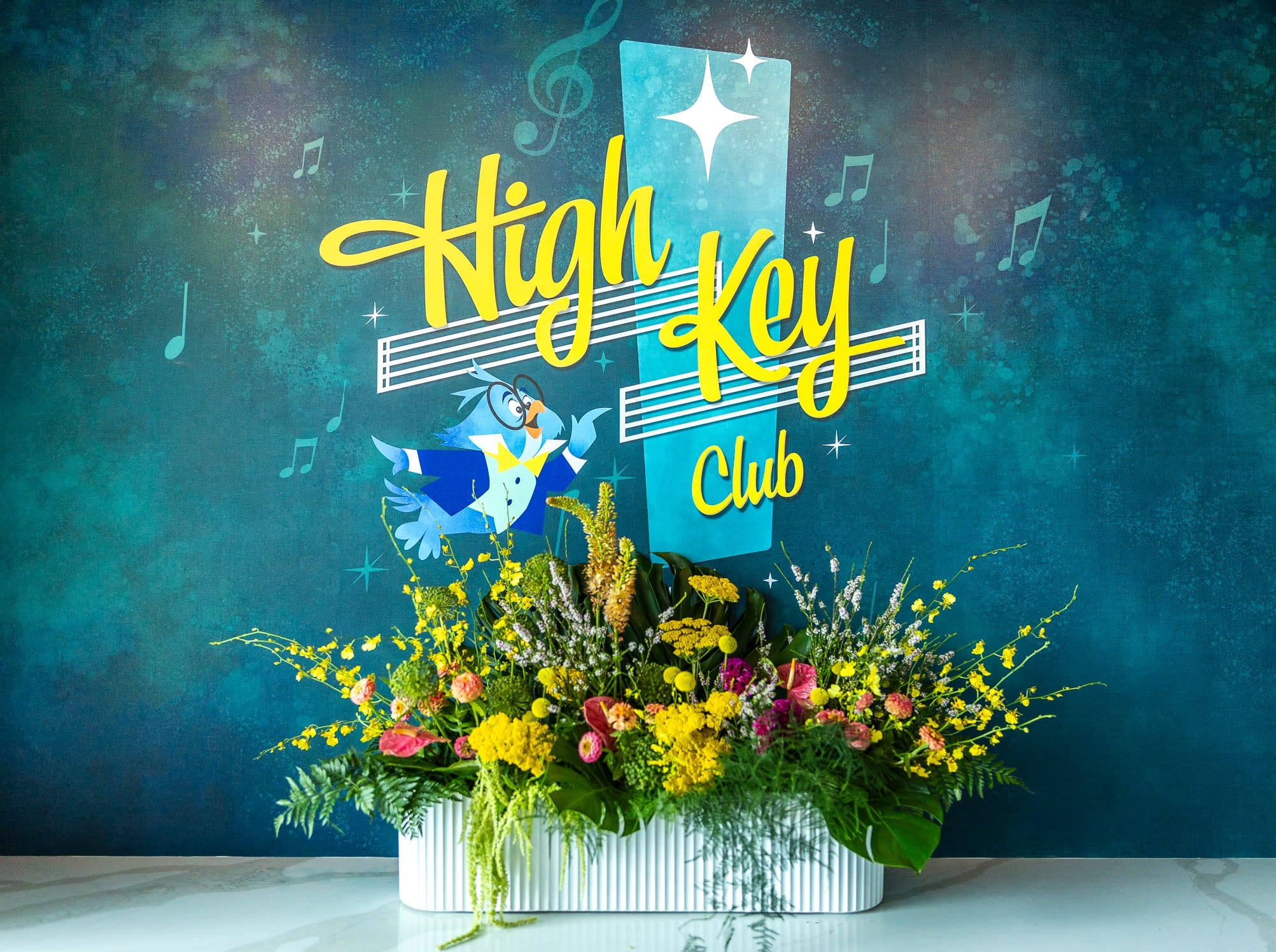 A colorful flower arrangement in front of the Disneyland Hotel’s High Key Club sign with musical notes and a cartoon bird.