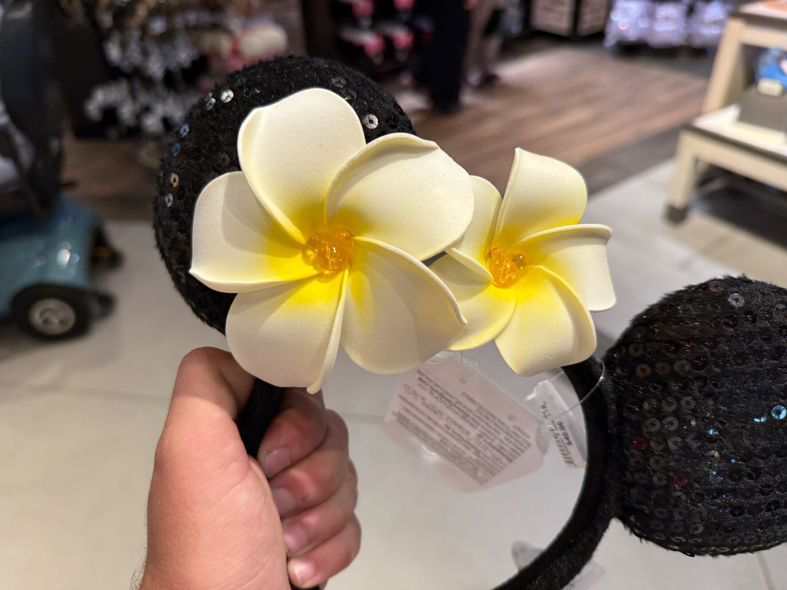A hand holds black sequin mouse ears with two large white and yellow plumeria flowers attached.