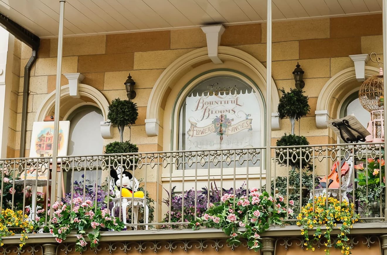 A balcony with flowers, greenery, and a Mickey Mouse statue in front of windows reading “Bountiful Blooms Flower & Garden.”