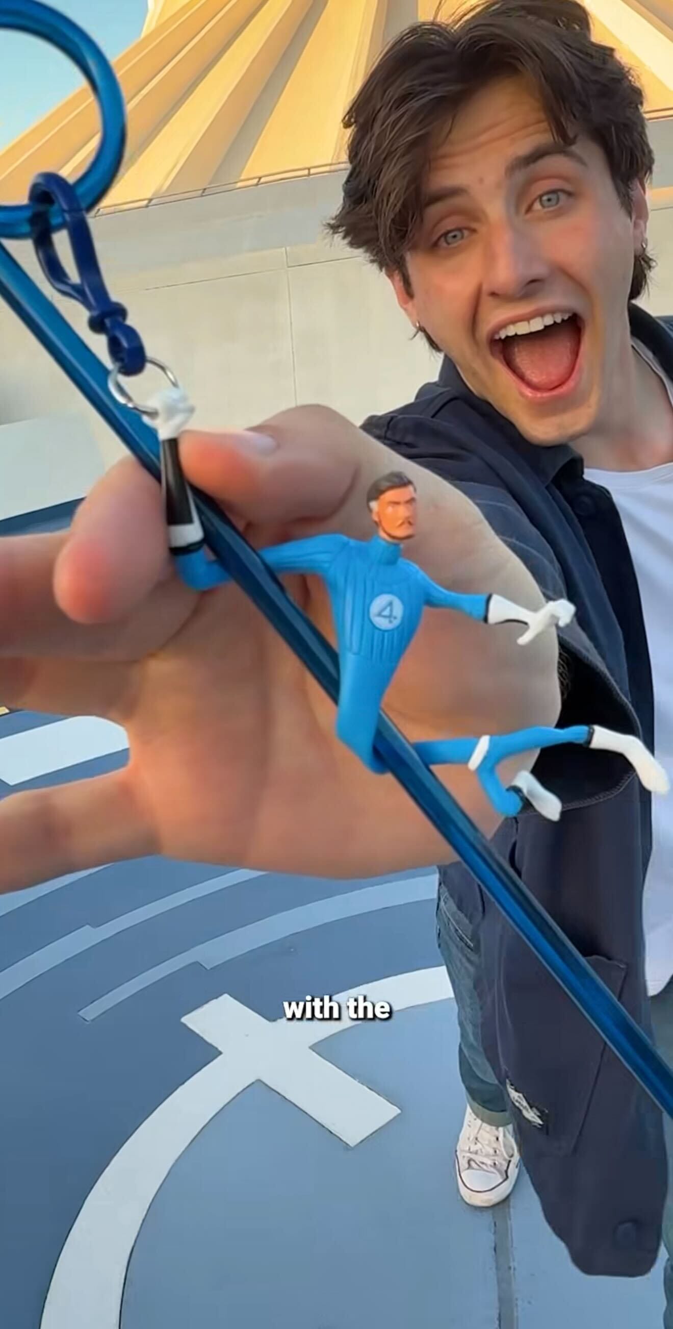 A smiling young man holds a blue Mister Fantastic keychain toward the camera with a building at Disneyland Resort behind him.