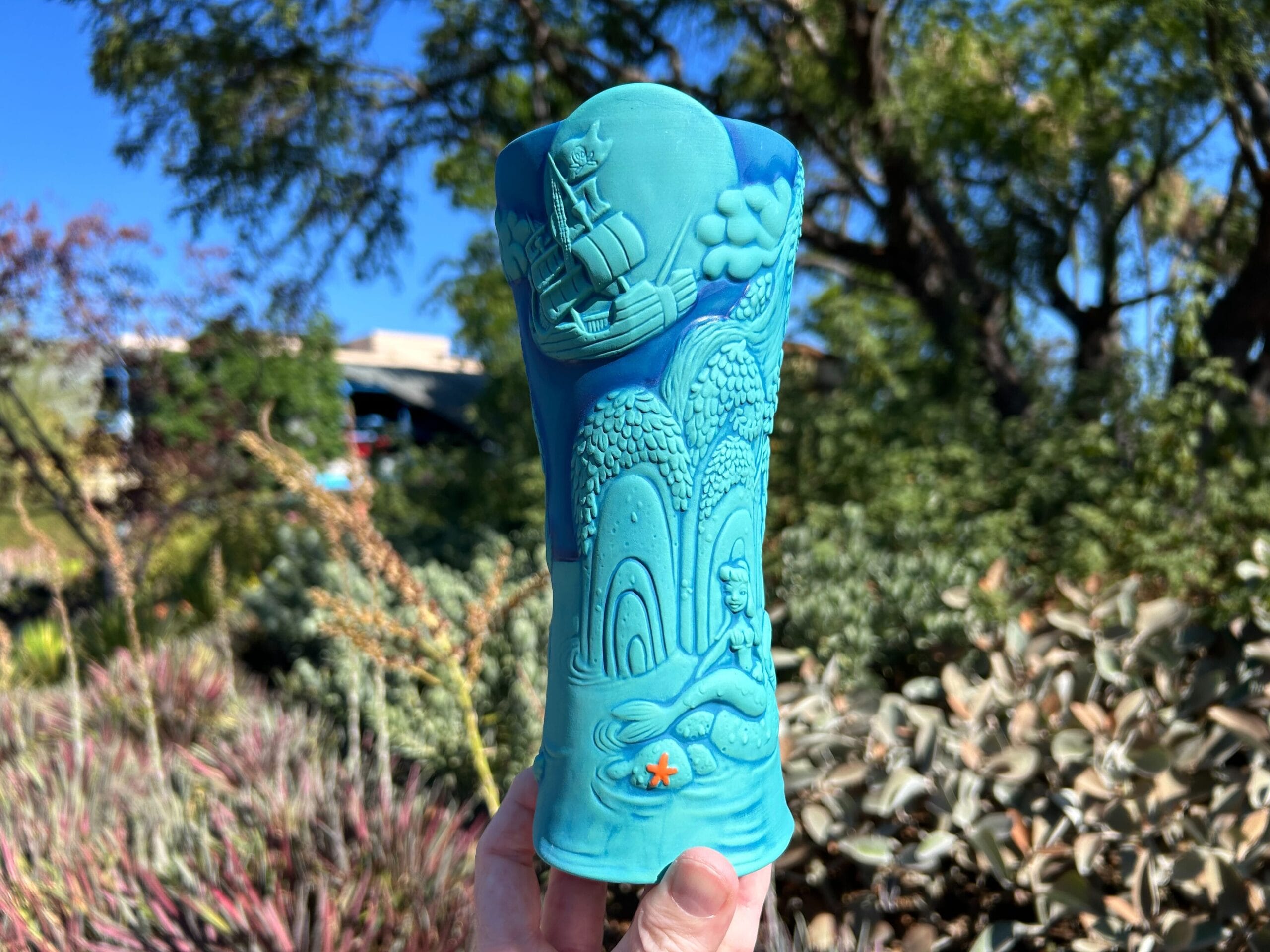 A turquoise mug with carved tropical designs, held outdoors among green, leafy plants and under a bright blue sky.