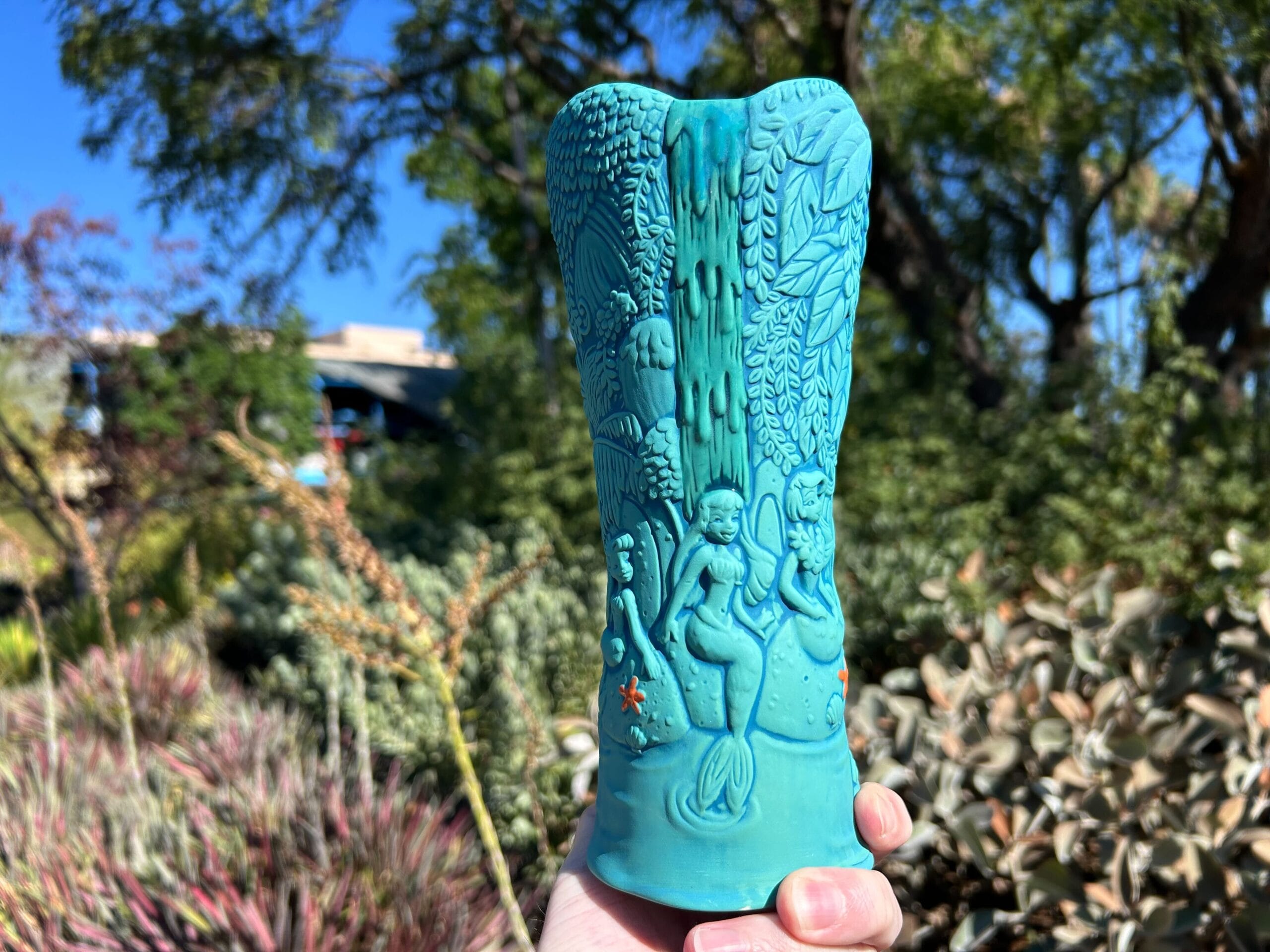 A hand holds a turquoise vase with mermaid carvings, lush greenery and blue sky in the background.