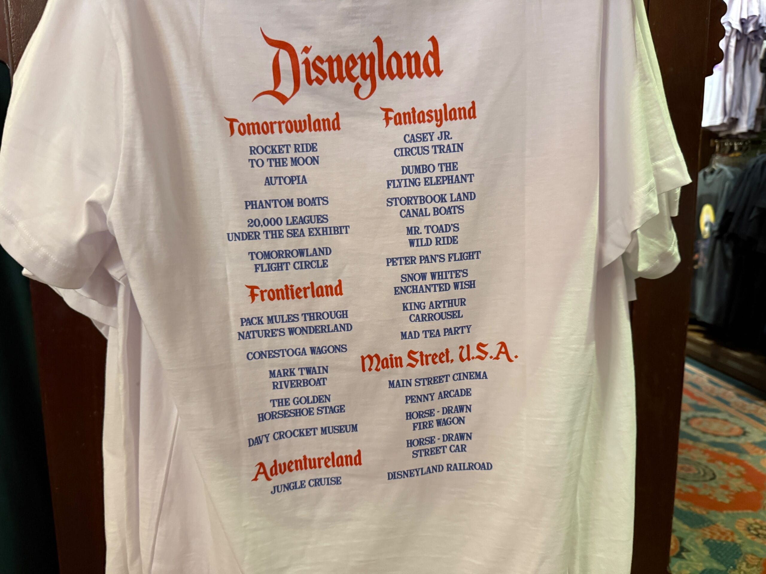 White Disneyland T-shirt listing classic attractions by area in red and blue text, displayed in a store.