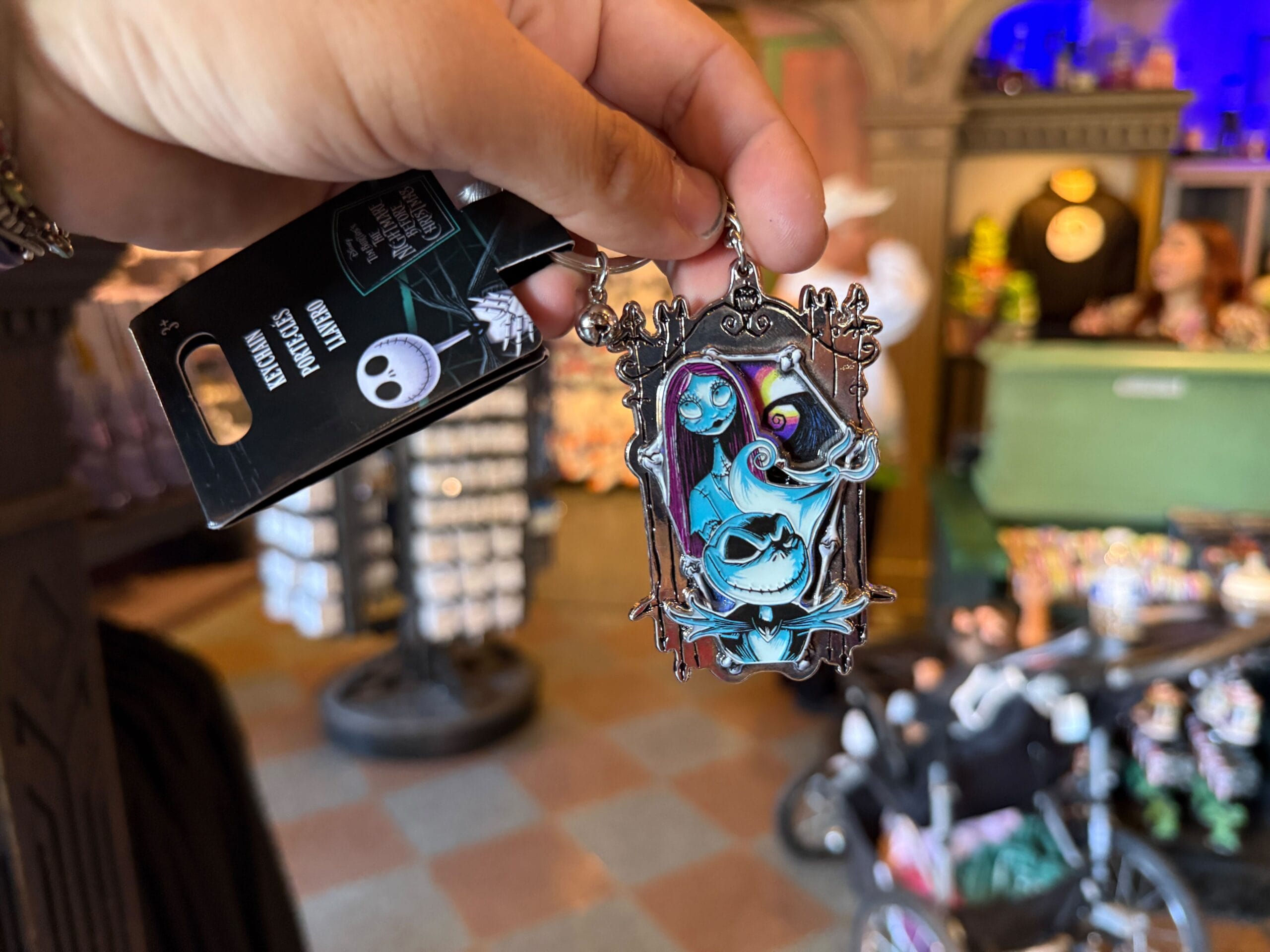 A hand holds Pumpkin King merchandise—a Nightmare Before Christmas keychain with Sally and Zero in a shop.