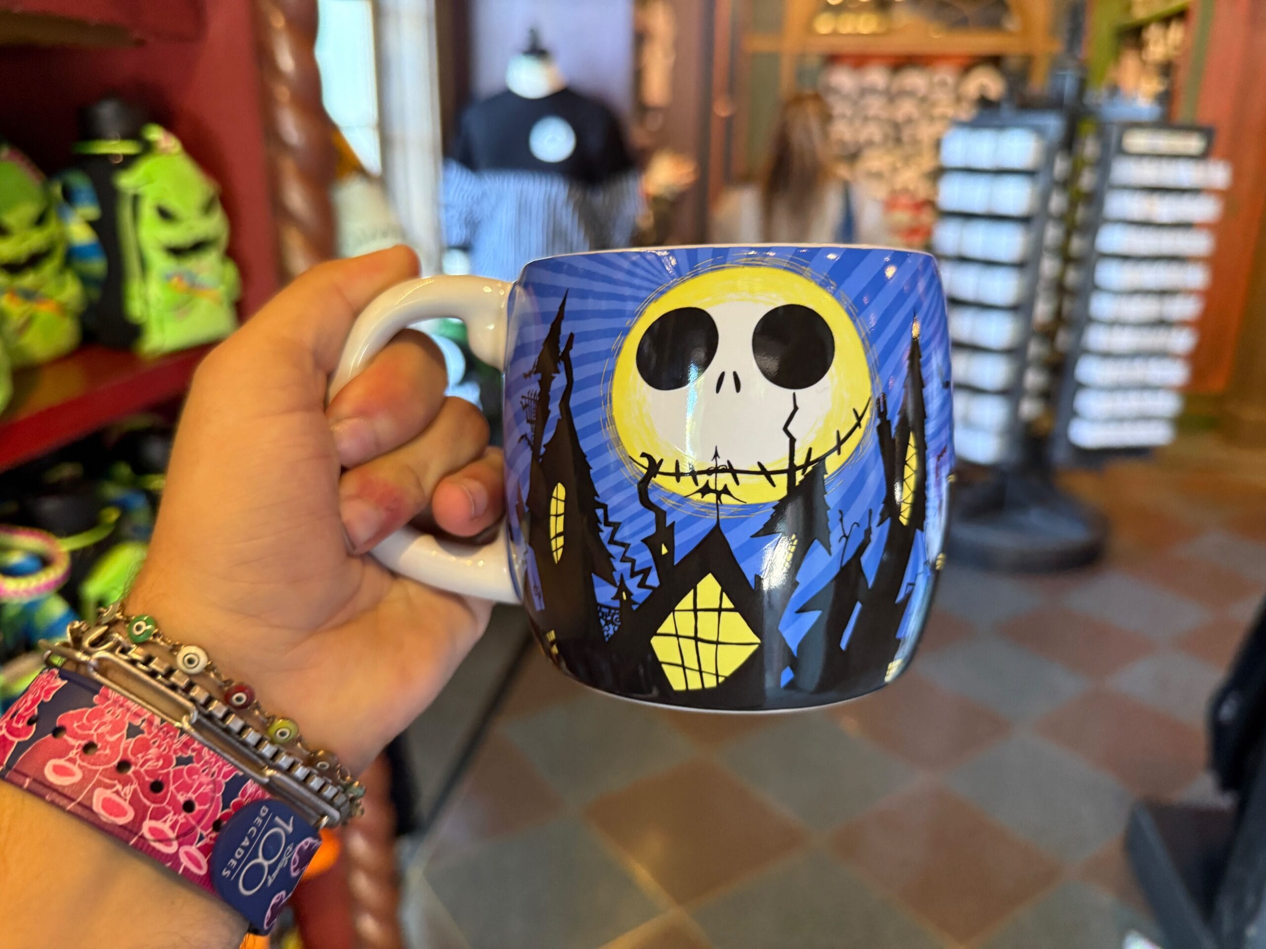 A hand holds a mug featuring the Pumpkin King, Jack Skellington, and a moonlit spooky scene from "The Nightmare Before Christmas.