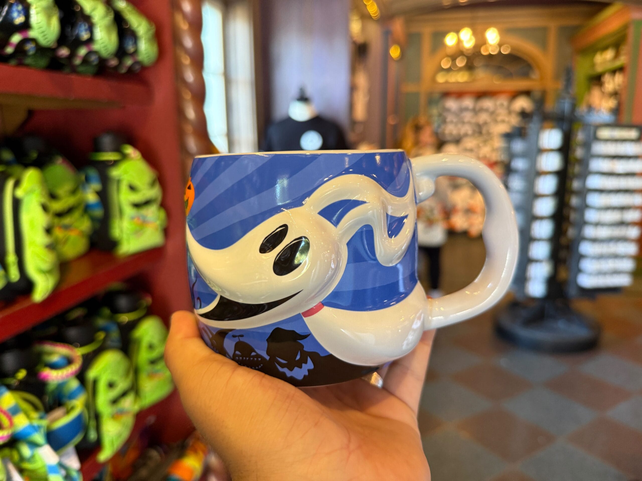 A hand holds a blue mug featuring Zero from The Nightmare Before Christmas among Pumpkin King merchandise in a colorful store.