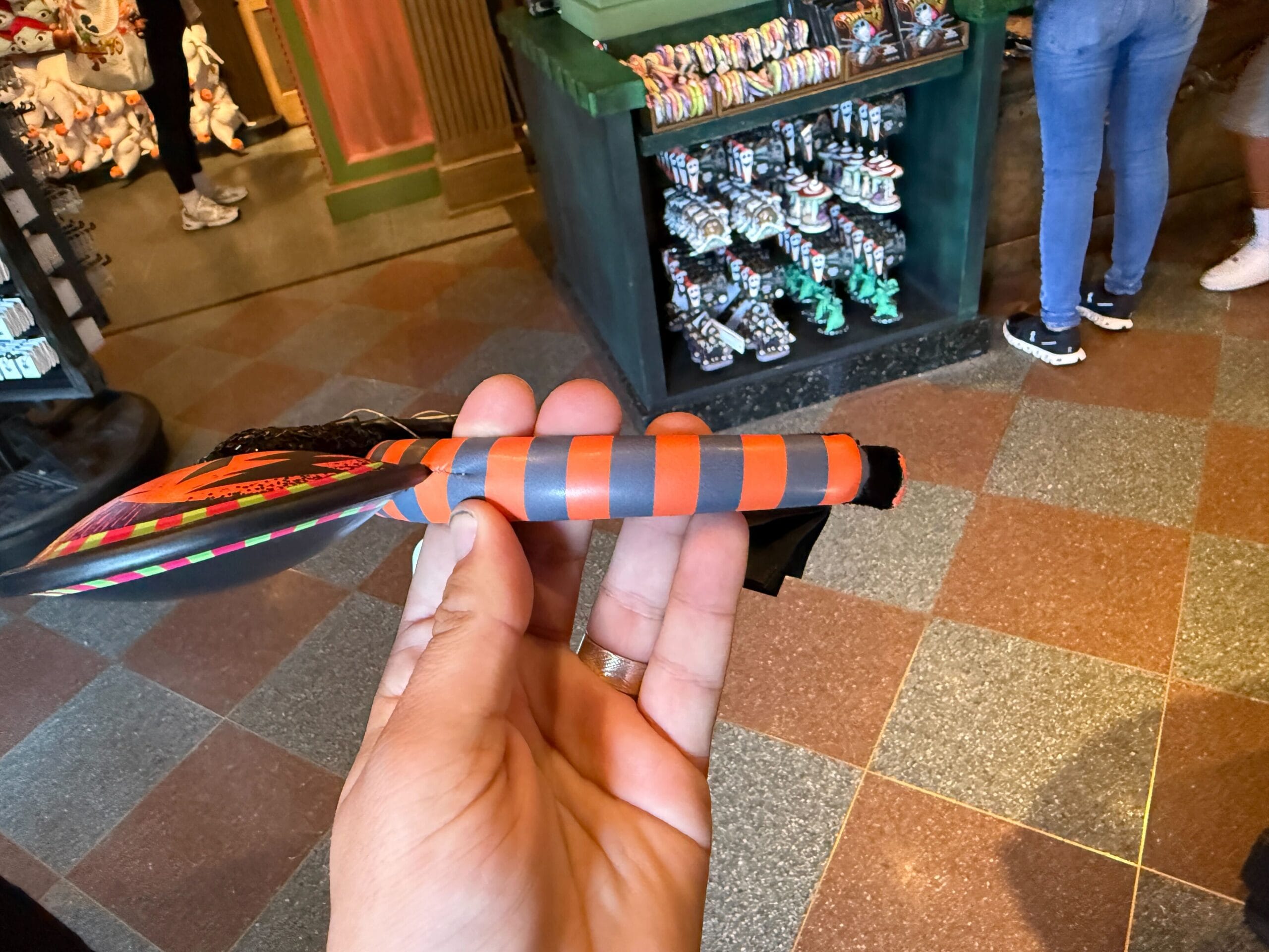 Hand holding a striped orange and gray toy knife, surrounded by Nightmare Before Christmas souvenirs in a store.