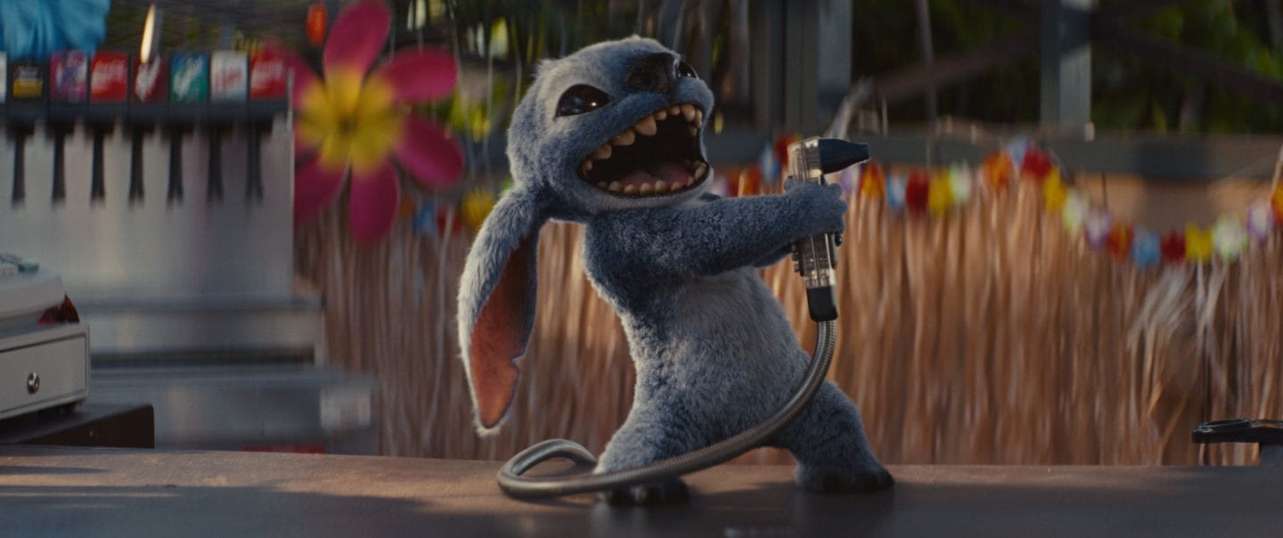 A blue alien from Lilo & Stitch stands on a counter, excitedly holding a soda fountain nozzle with its mouth open wide.