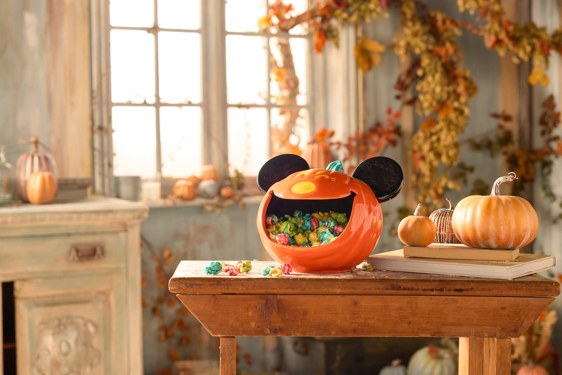 A Mickey Mouse pumpkin candy bowl from the Halloween Collection sits on a wooden table in an autumn-themed room.