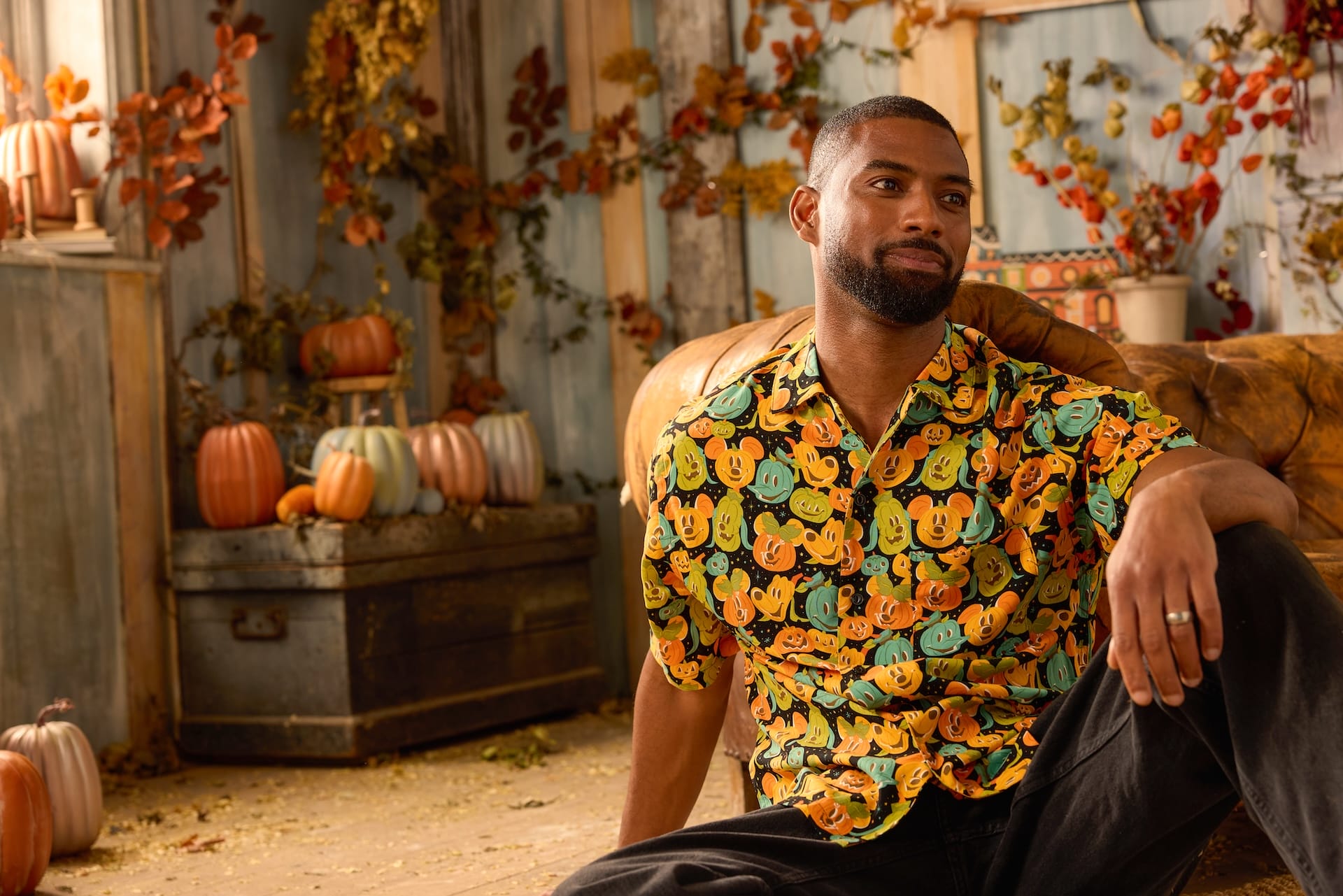 Man in a colorful pumpkin shirt sits in an autumn room with pumpkins, fall decor, and Disney Store Halloween Collection accents.