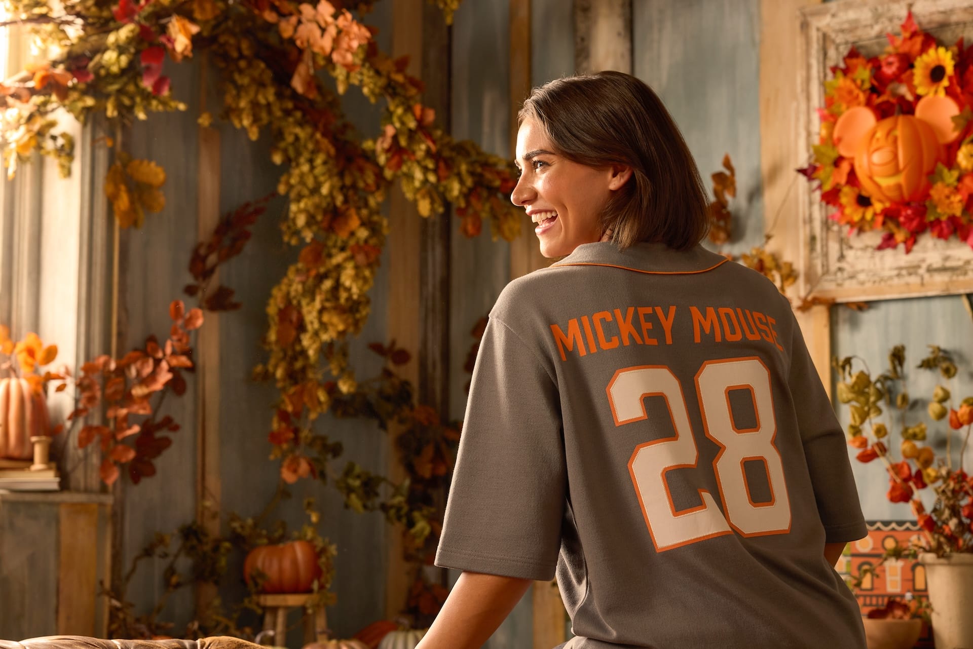 A smiling woman in a "Mickey Mouse 28" shirt stands in a room with pumpkins from the Halloween Collection.