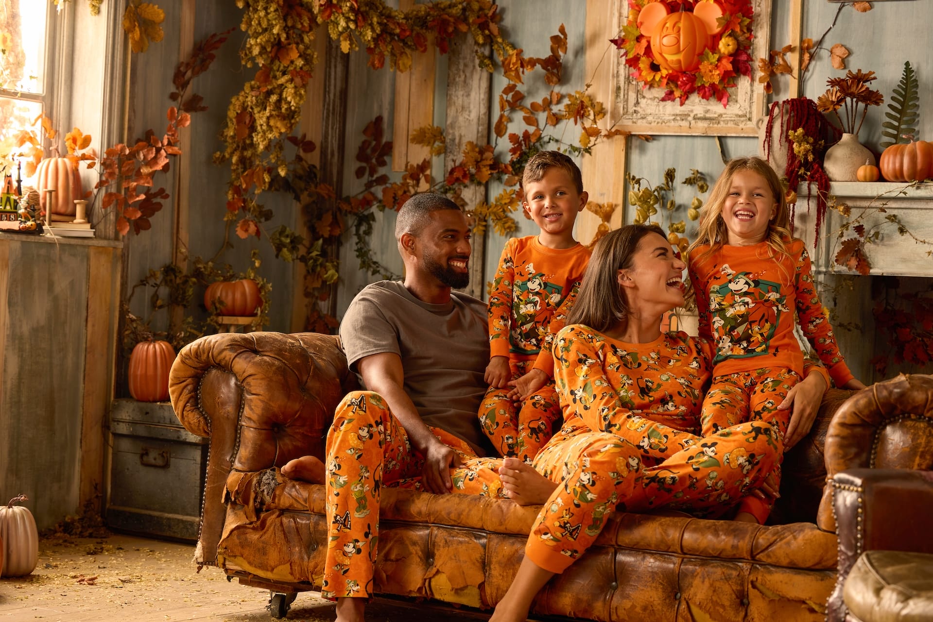 A family in matching orange pajamas from the Halloween Collection smiles on a couch with autumn leaves and pumpkins.