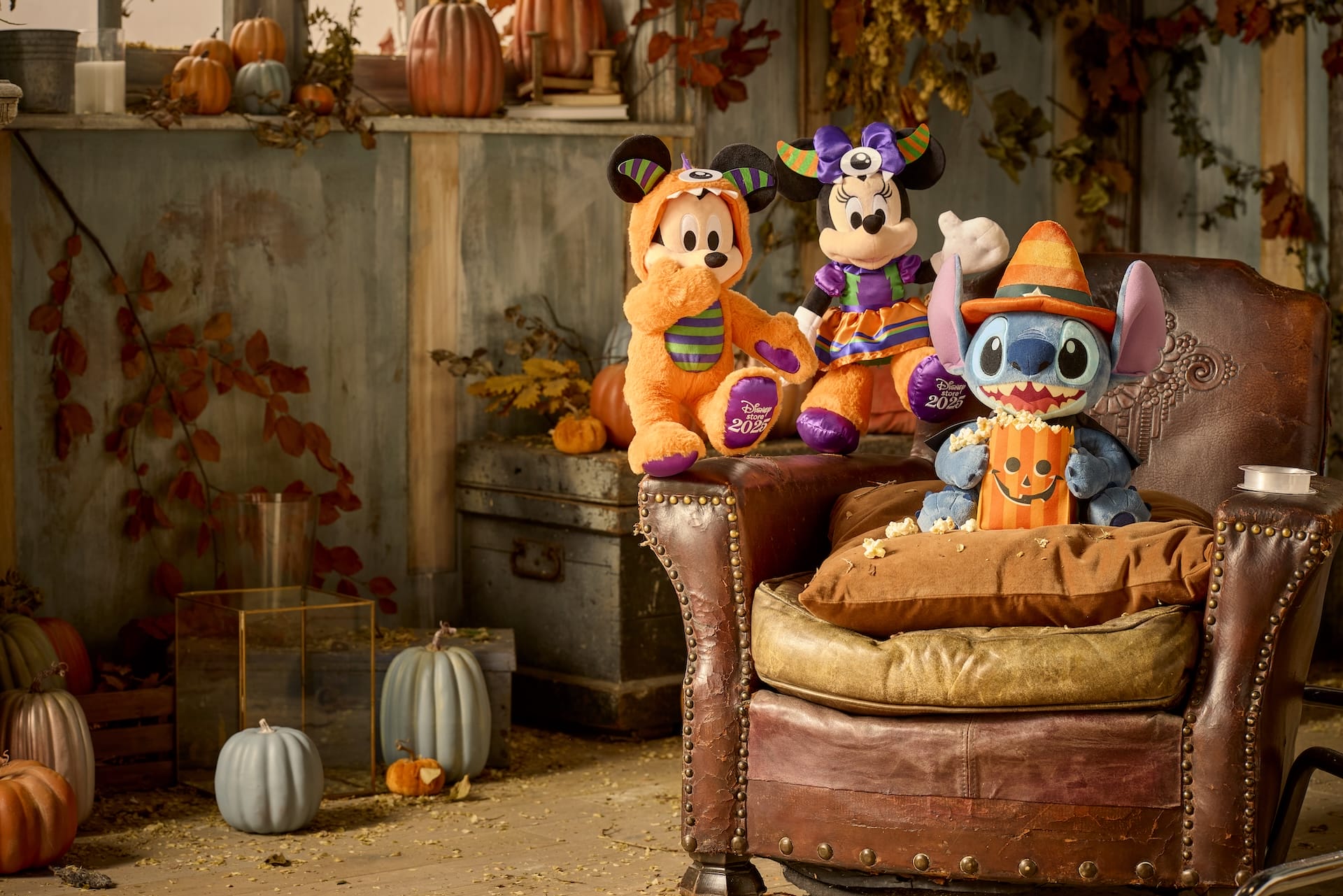 Plush toys in Halloween costumes sit on a leather chair and table, surrounded by pumpkins and autumn decor.