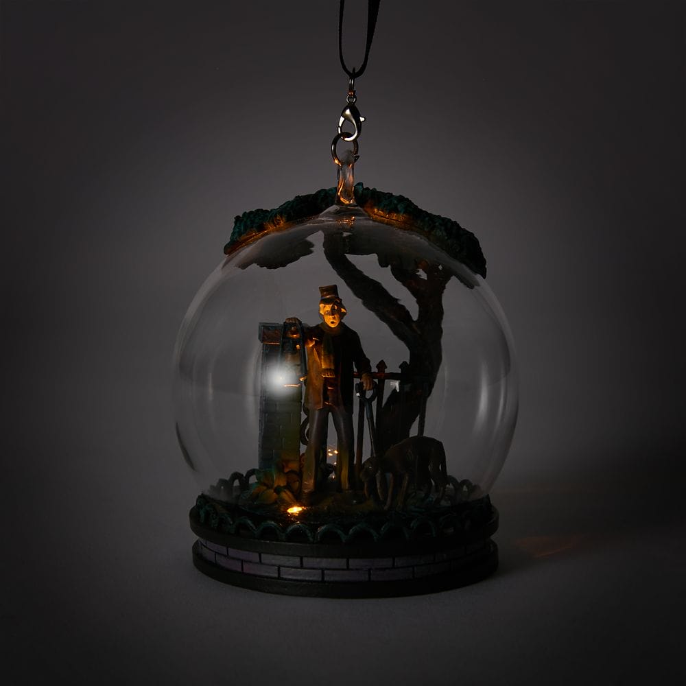 A glass ornament from the Haunted Mansion Collection, featuring a man with a lantern by a tree, glows inside the globe.