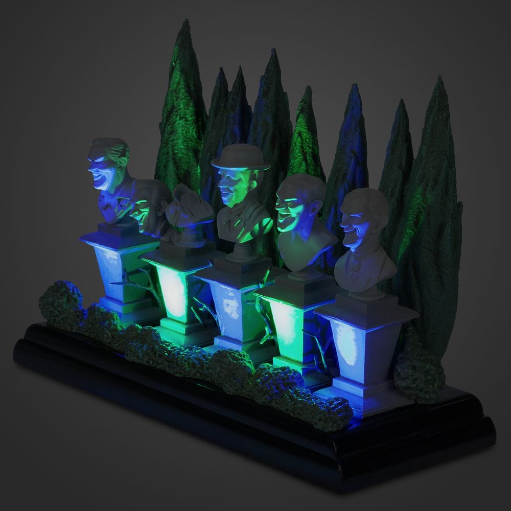 Five sculpted busts on pedestals glow with green and blue lights amid trees—an eerie Haunted Mansion Collection scene.