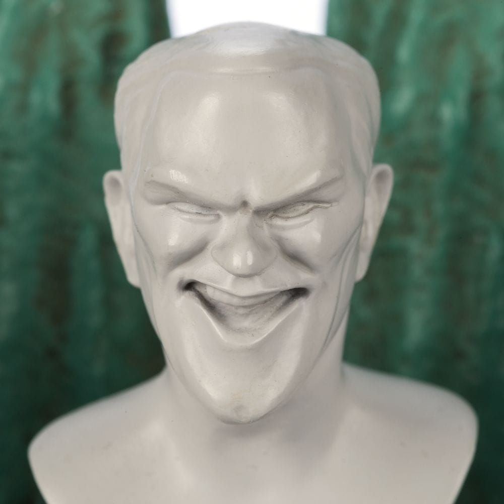 A white sculpture bust with an exaggerated grin and sharp features, from the Haunted Mansion Collection, against a green background.