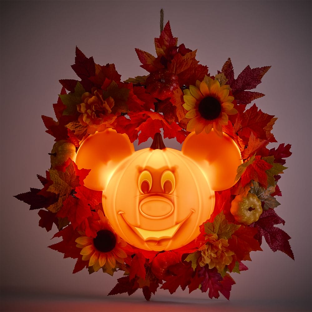 A glowing pumpkin shaped like Mickey Mouse is surrounded by autumn leaves, sunflowers, and small pumpkins.