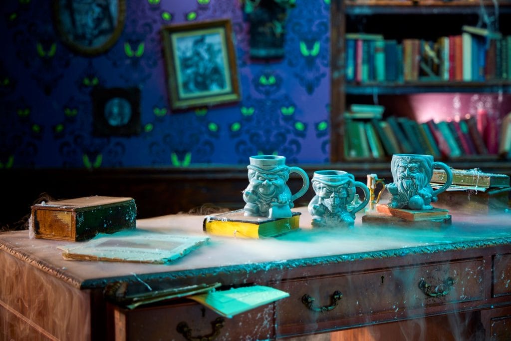 Three ornate mugs sit on a cobwebbed desk in a spooky, haunted mansion library, like a scene from the Disney Store.