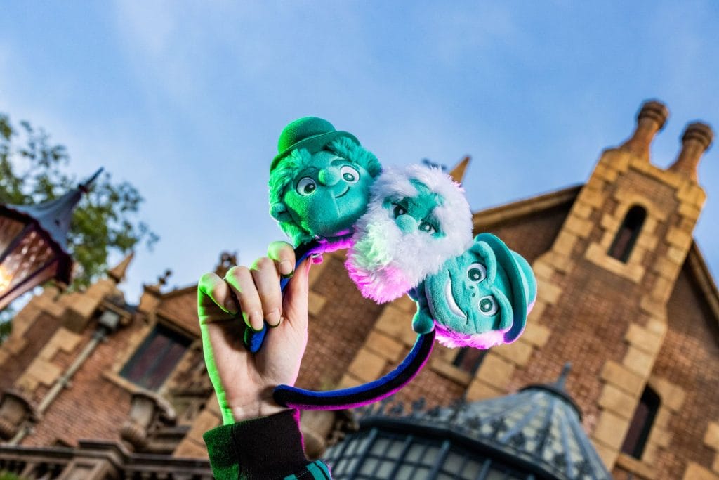 Hand holding a Haunted Mansion headband with three ghost faces in front of a Disney Store brick building.
