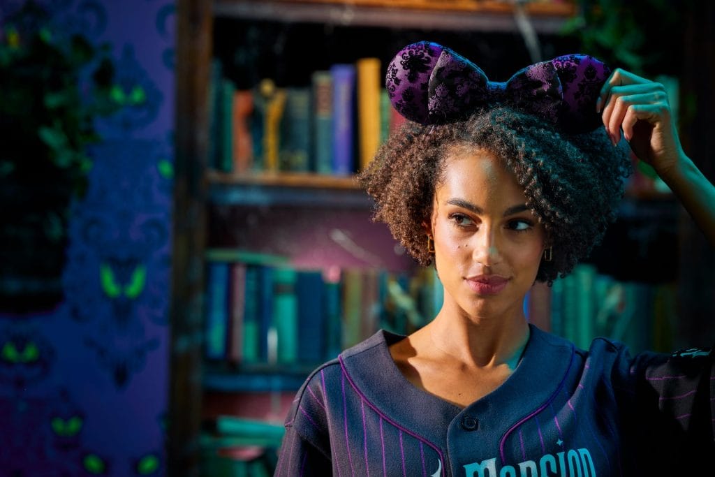 Woman with curly hair in purple mouse ears, Disney Store vibe, stands by bookshelf, looking to the side and smiling softly.