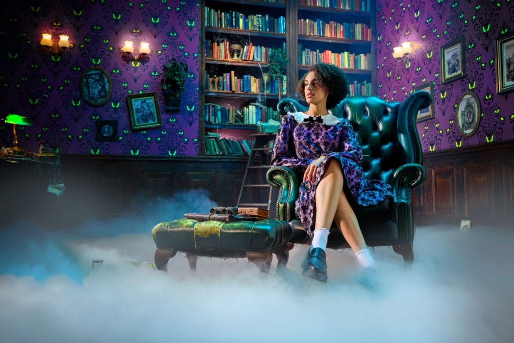A woman in a vintage dress sits in a green armchair in a foggy, haunted mansion library with purple wallpaper.