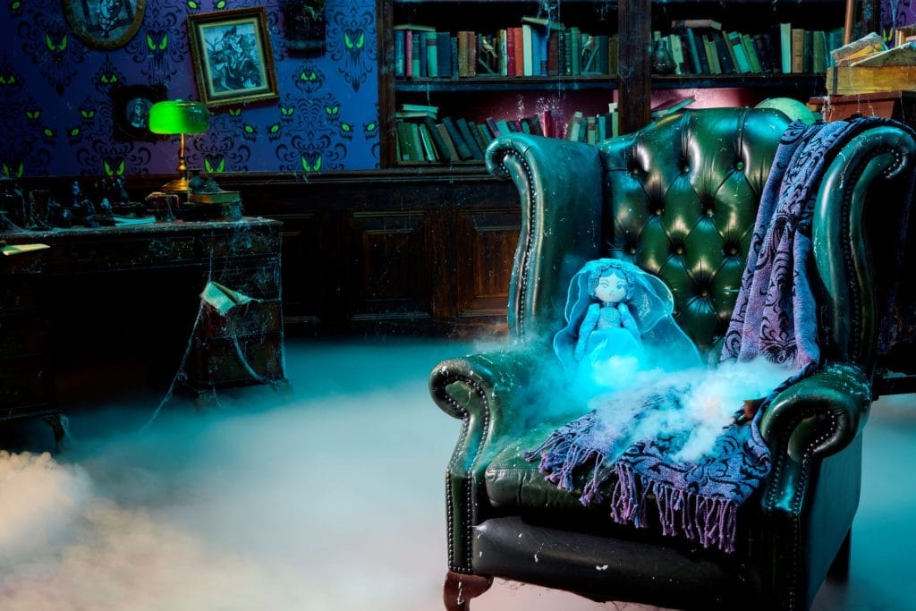 A haunted mansion room with a foggy floor, eerie lighting, an armchair, blue doll, and bookshelves.