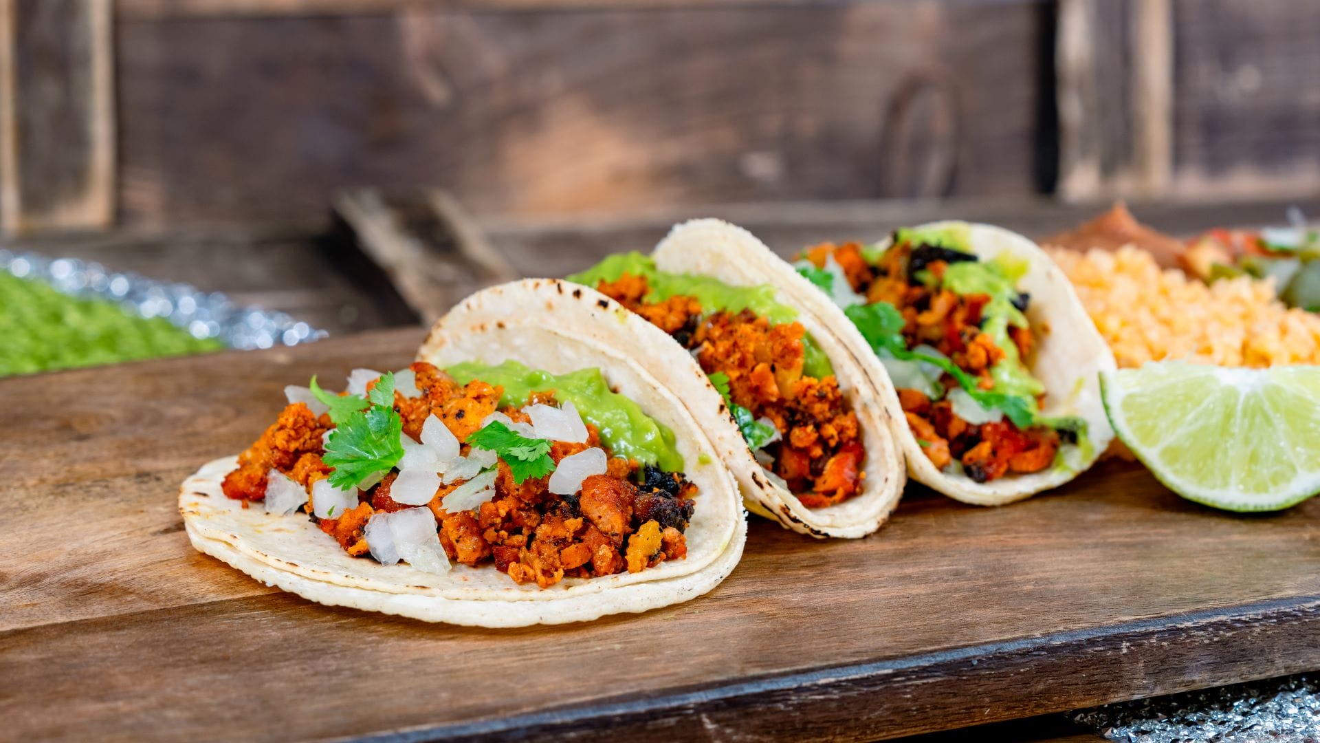 Three tacos from the 2025 menu at Plaza de la Familia, topped with onions, cilantro, lime wedges, and rice.