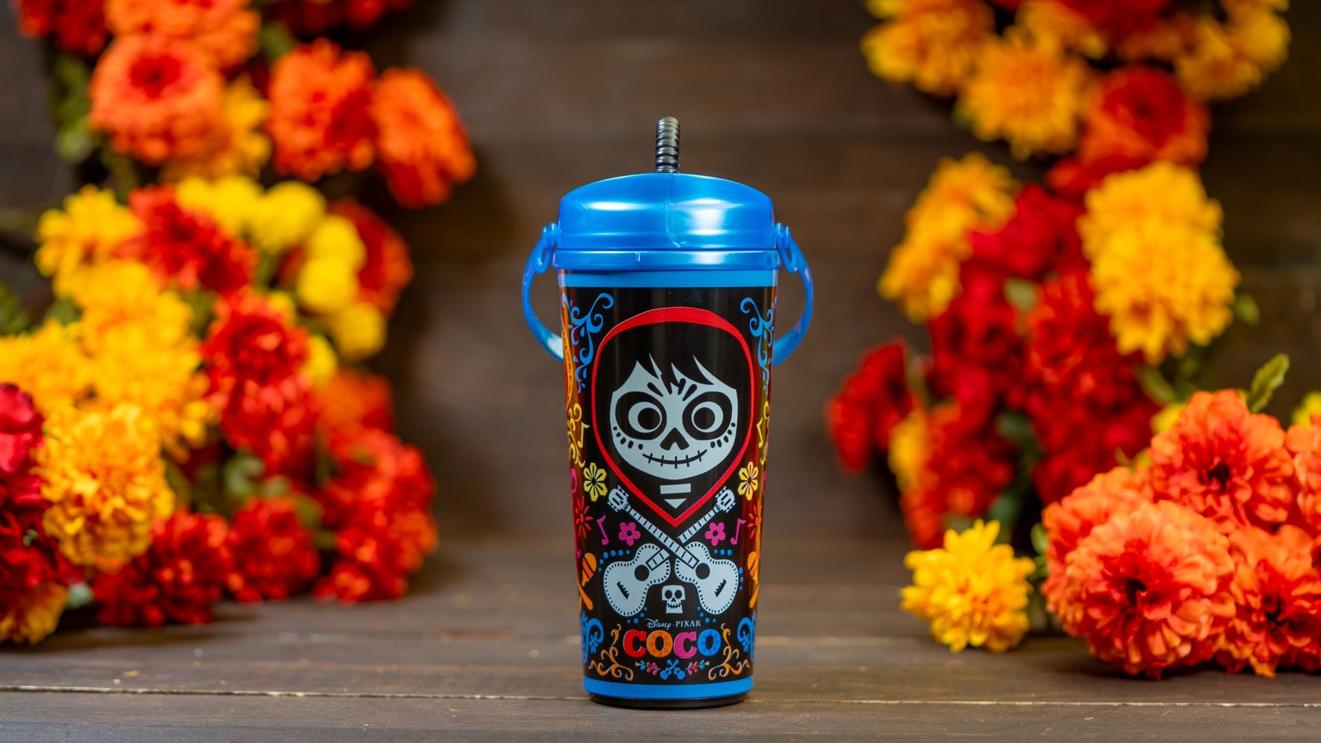 A colorful Coco Sipper cup with a blue lid, surrounded by vibrant marigolds, inspired by Disneyland Resort.