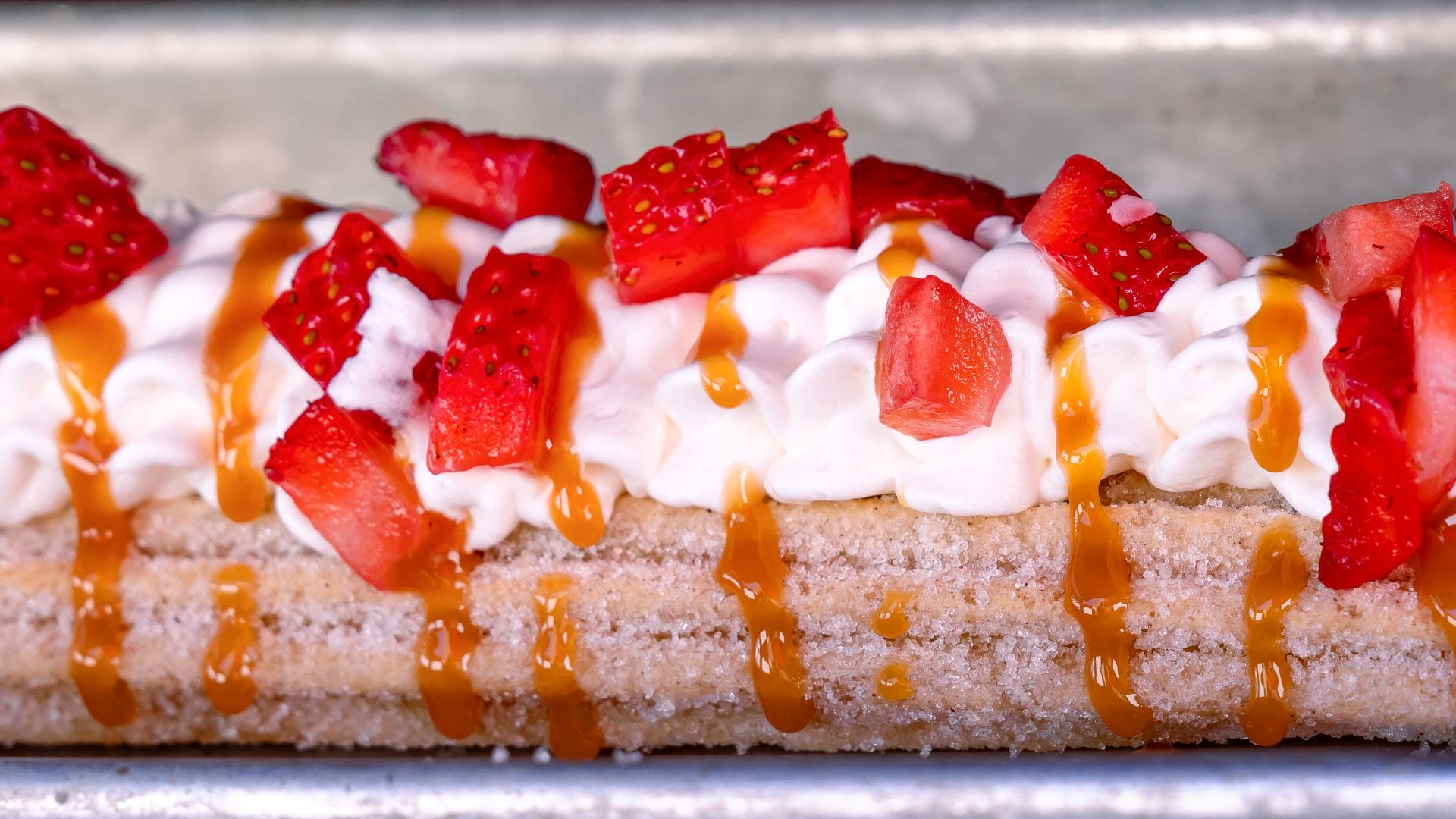 A sugar-coated churro topped with whipped cream, fresh strawberries, and caramel—part of Plaza de la Familia 2025 menu items.