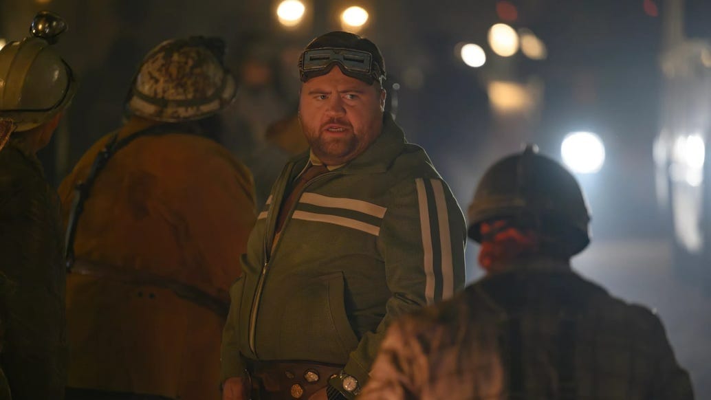A man in a green jacket and goggles stands among workers in helmets, like the Fantastic Four taking First Steps at night.