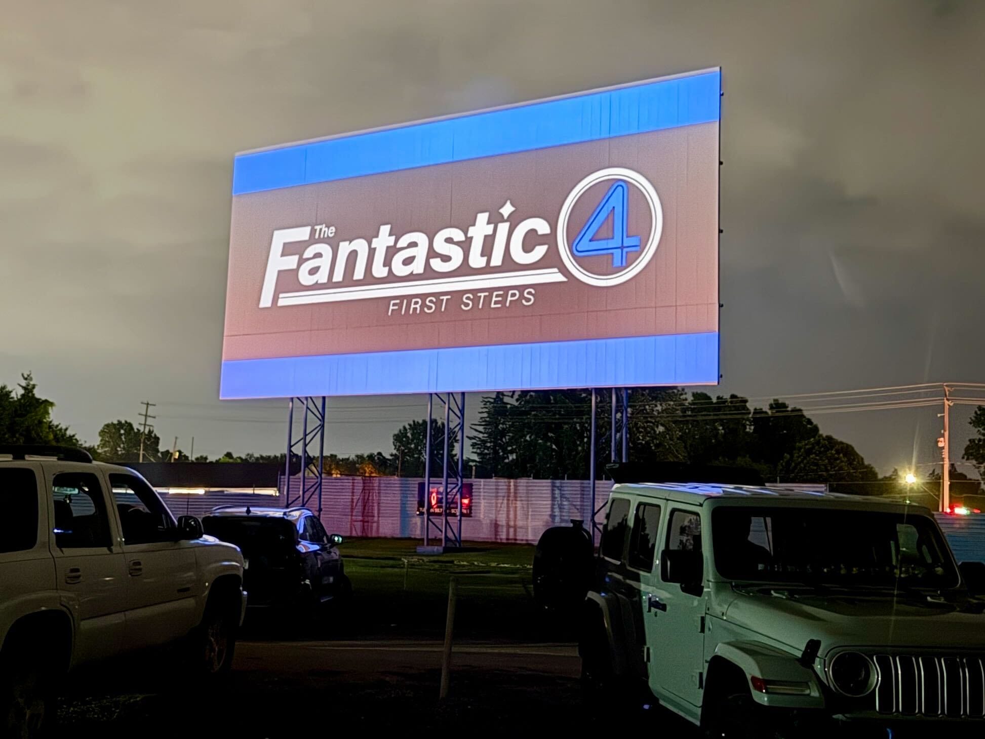 A drive-in movie screen shows "Fantastic Four: First Steps" at night, cars parked to watch Reed Richards’ debut.