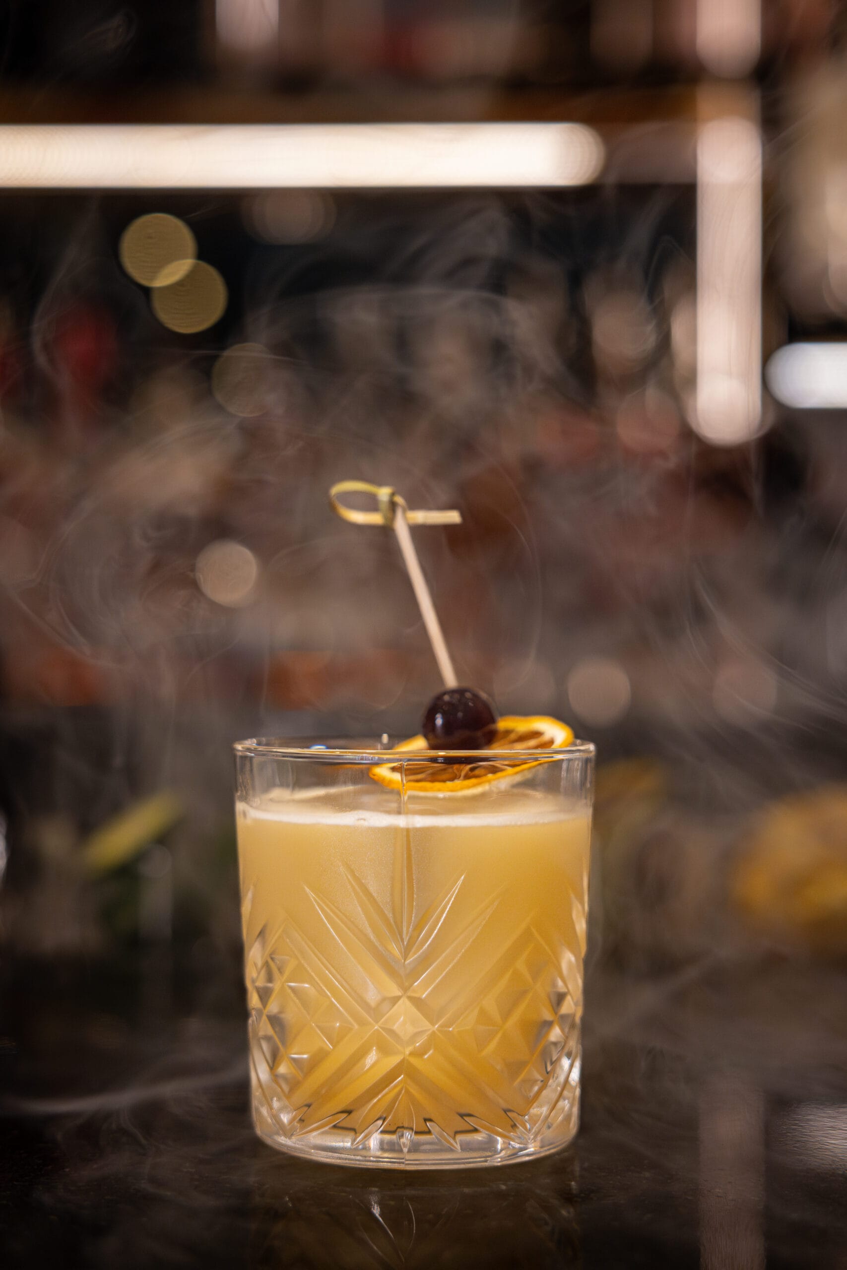 A cocktail in a patterned glass at Bourbon Steak, garnished with orange and cherry, set against a smoky background.