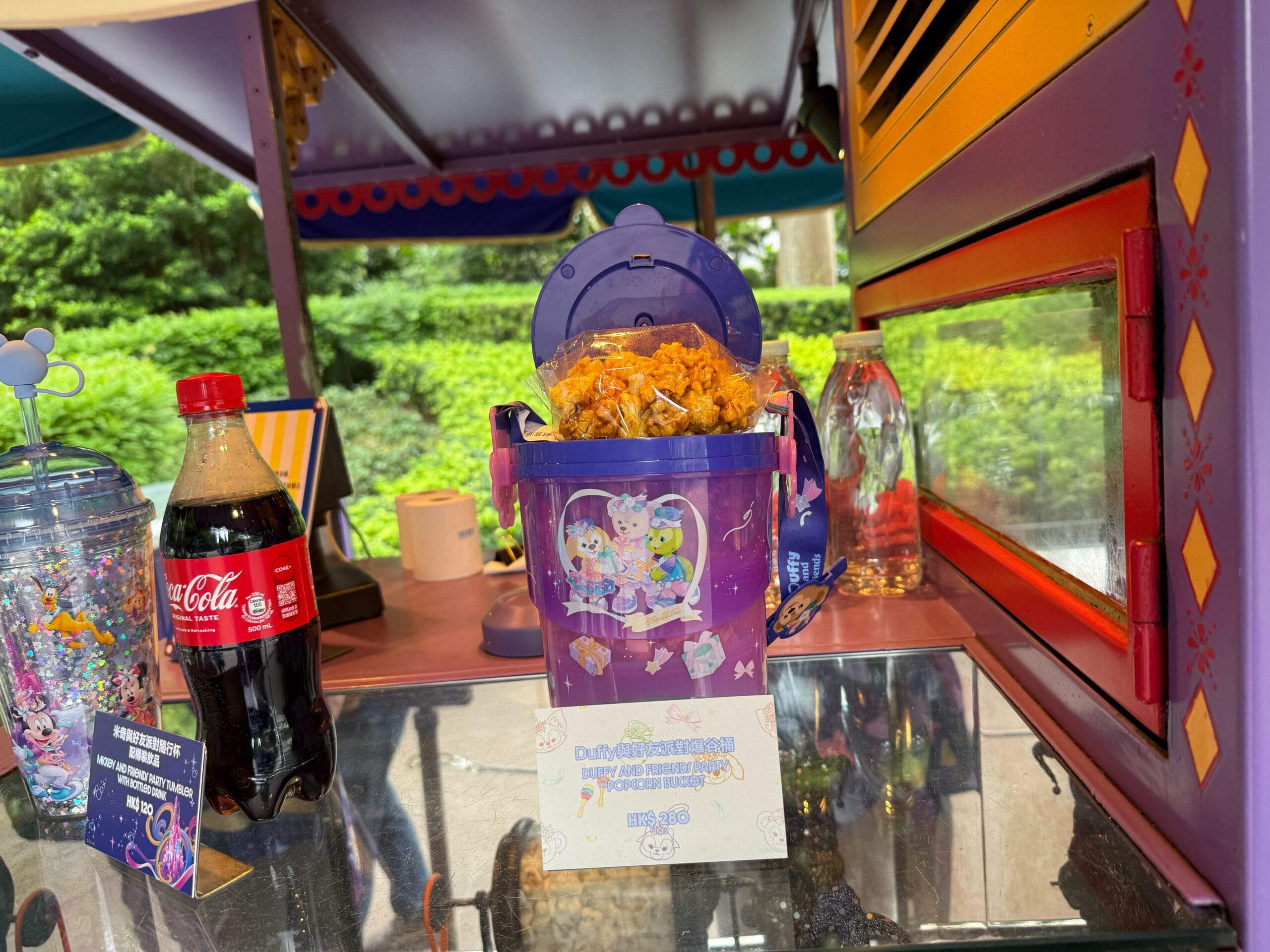 Purple popcorn bucket with cartoon characters, filled with popcorn, beside a Coke bottle on a colorful counter.