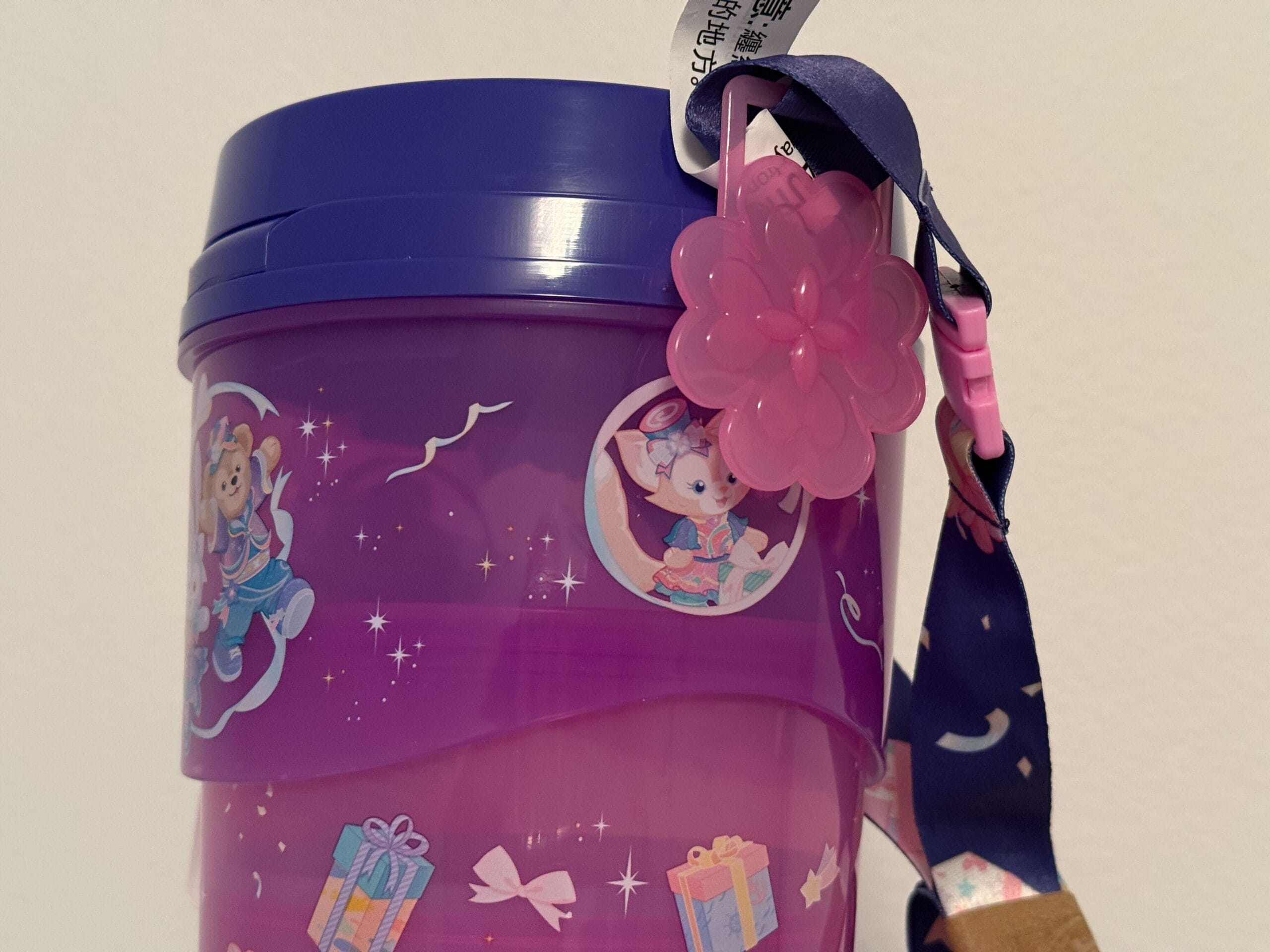 A purple cup with cartoon animals, presents, a pink flower charm, and a blue strap attached to the lid.